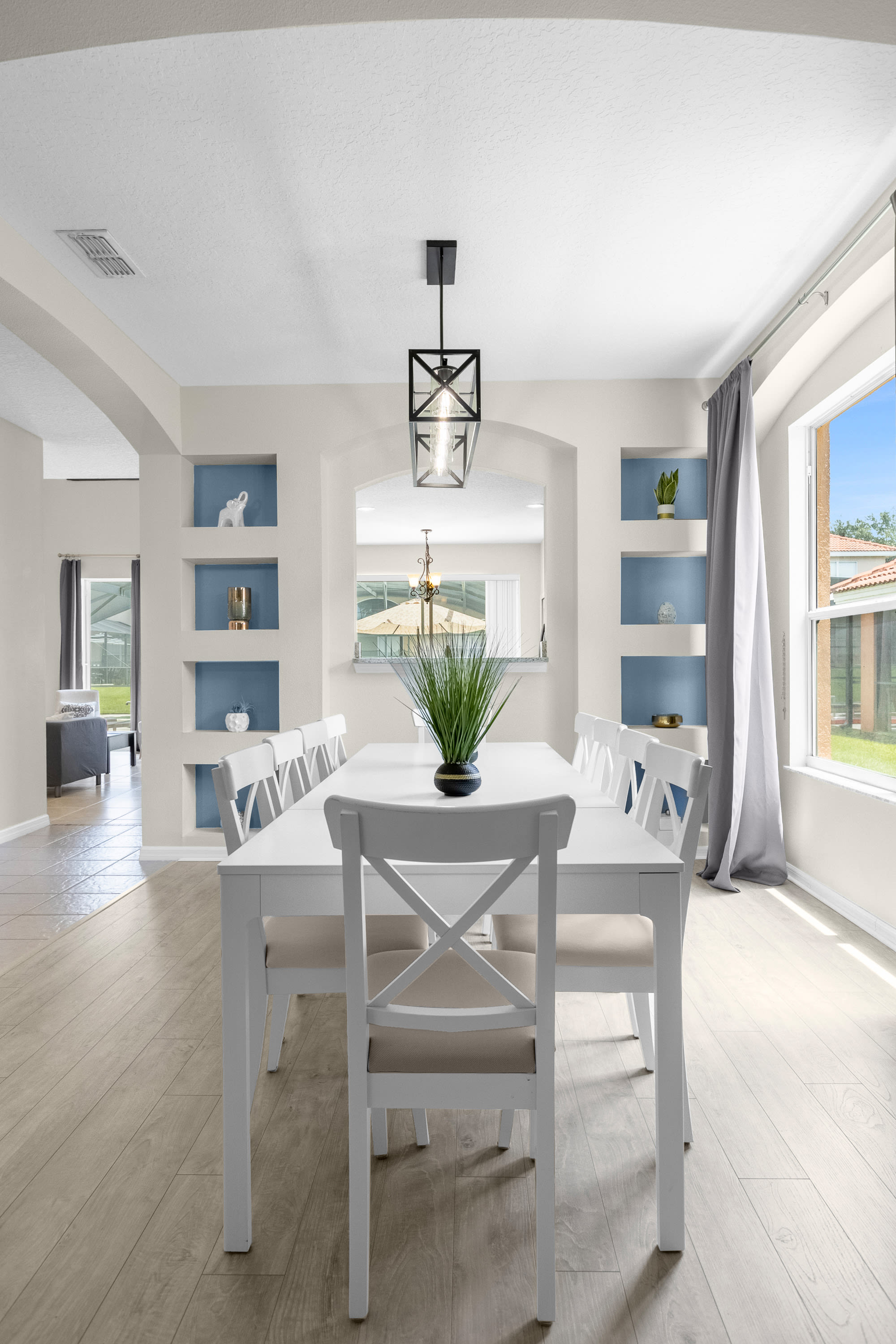 Formal dining room with light wood-look flooring, a long white dining table set for eight with white cross-back chairs, and a black linear chandelier. It features built-in blue display shelves and a large window with gray curtains.