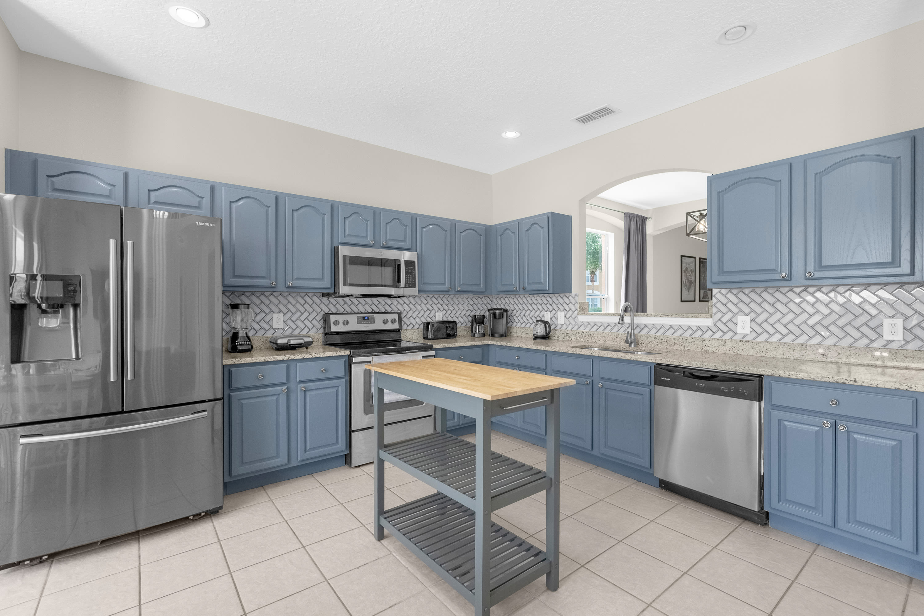Modern kitchen featuring light blue shaker-style cabinets, light granite countertops, a white herringbone subway tile backsplash, and stainless steel appliances including a refrigerator, range, microwave, and dishwasher, with a movable wooden island.