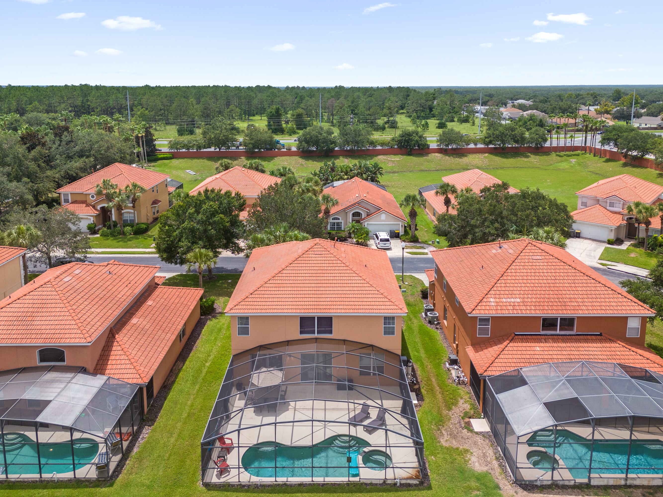 An aerial view of a residential neighborhood featuring several two-story homes with orange tiled roofs and screened-in private pools in their backyards. A lush green landscape and distant tree line frame the community under a clear sky.