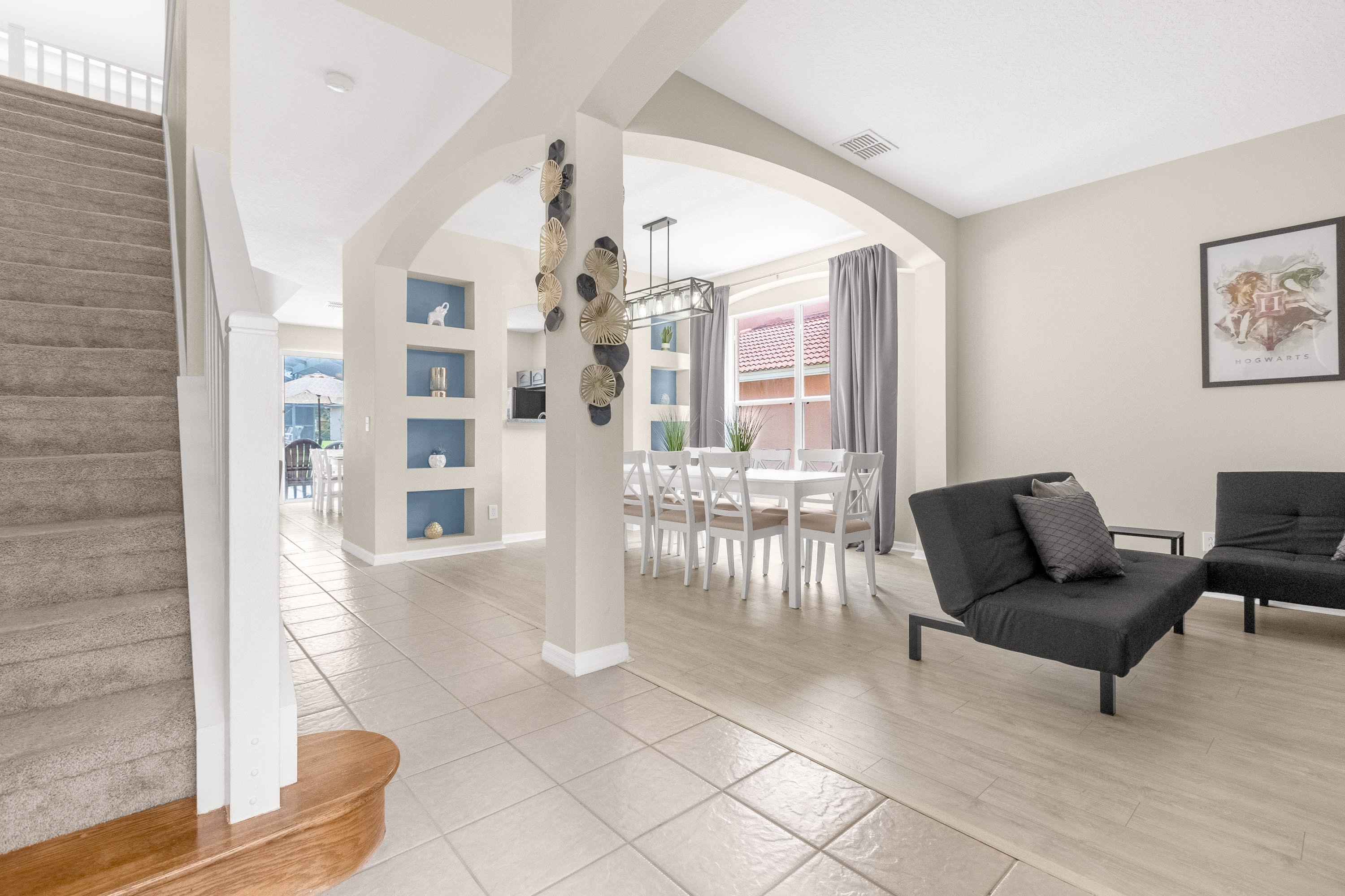 Entryway and open-concept living space with light tile and wood-look flooring, a staircase with white railings, and a dark gray futon. It leads to a dining area with a white table and built-in blue shelves.