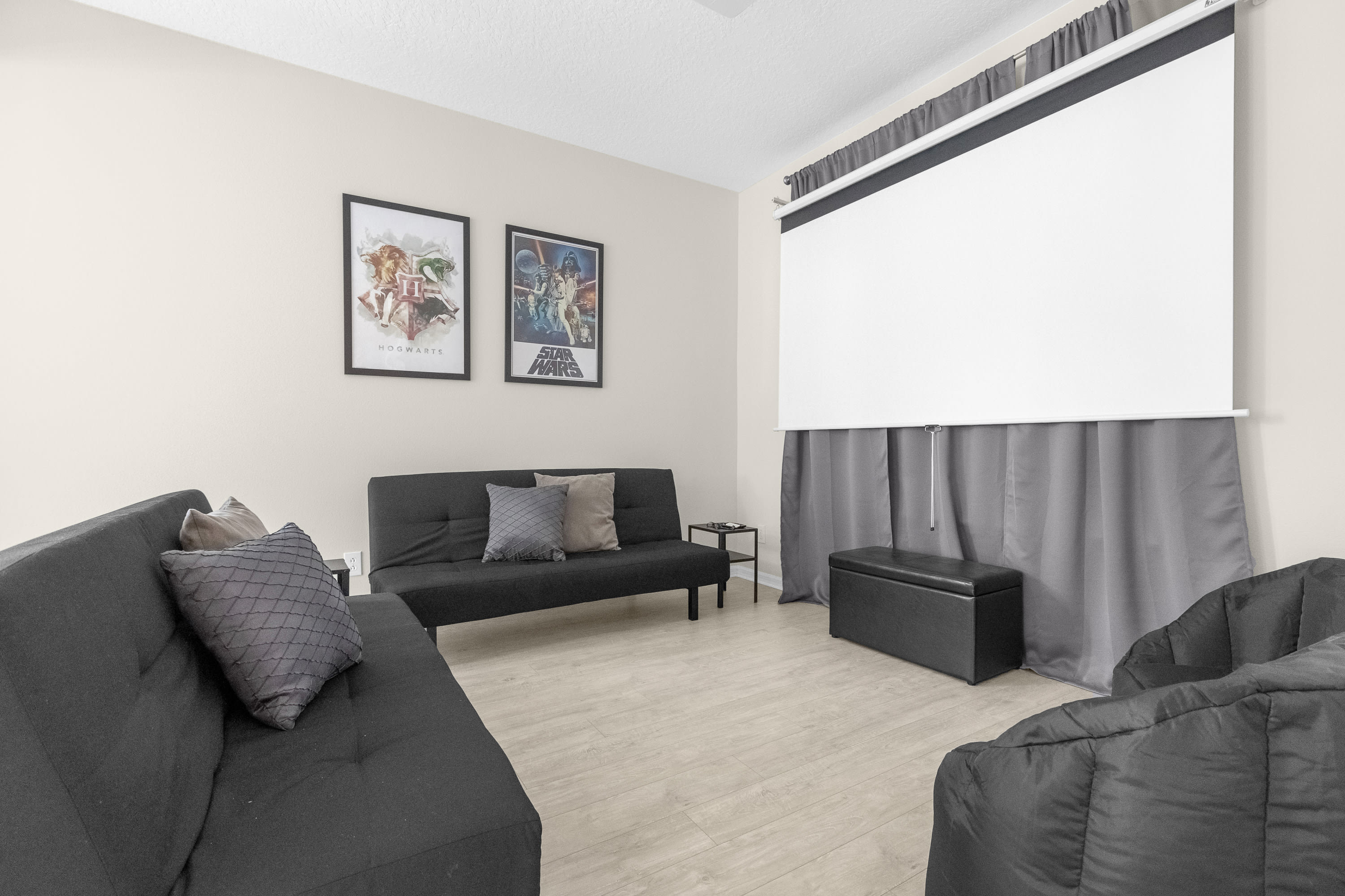 A cozy media room features two black futon sofas and bean bag chairs, facing a large pull-down projector screen. The light-colored walls are adorned with "Hogwarts" and "Star Wars" movie posters, and the room has light wood-look flooring.