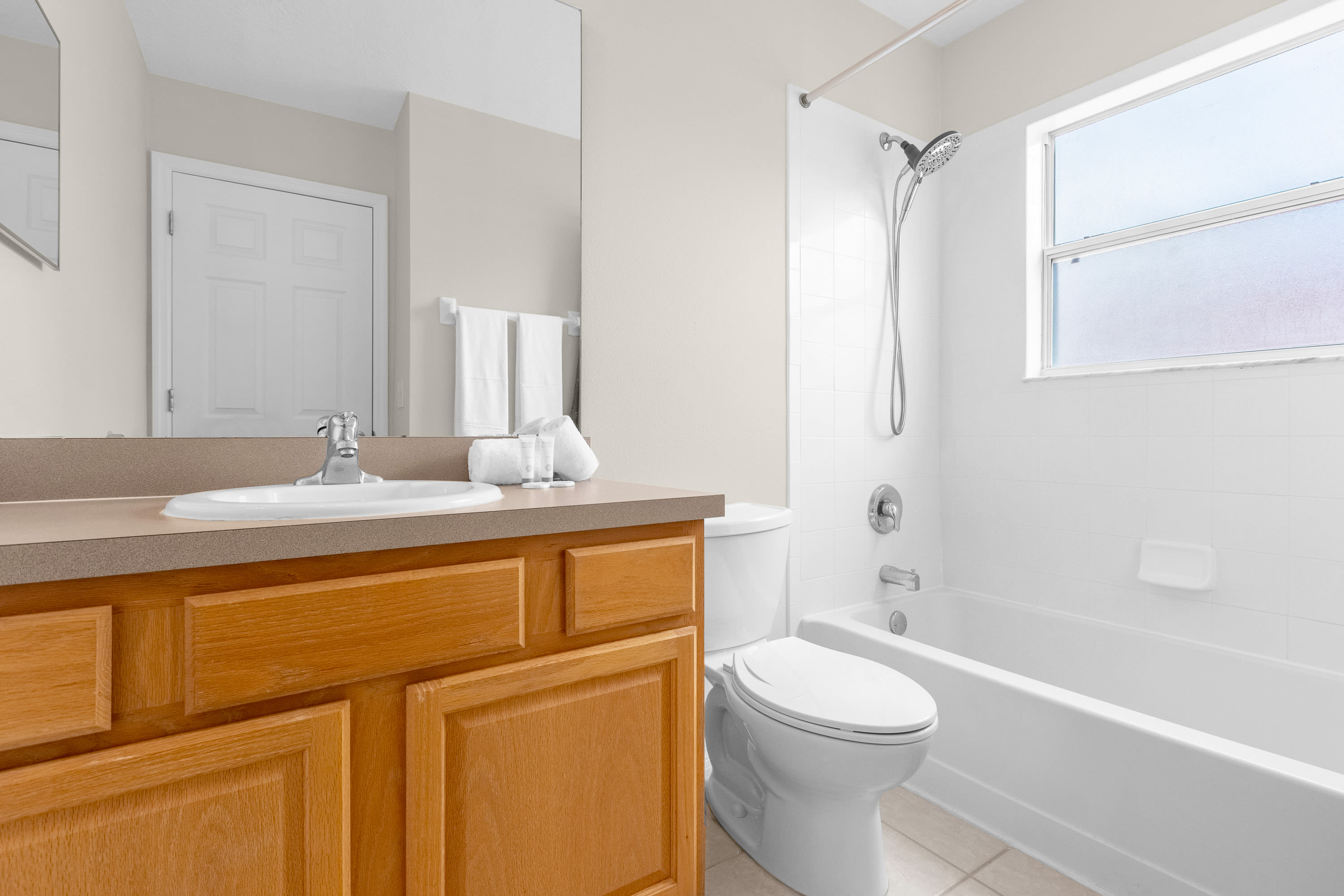 A clean bathroom with a light wood vanity, single sink, and a large mirror, positioned next to a white toilet and a shower-tub combo with white tiled walls and a frosted window.