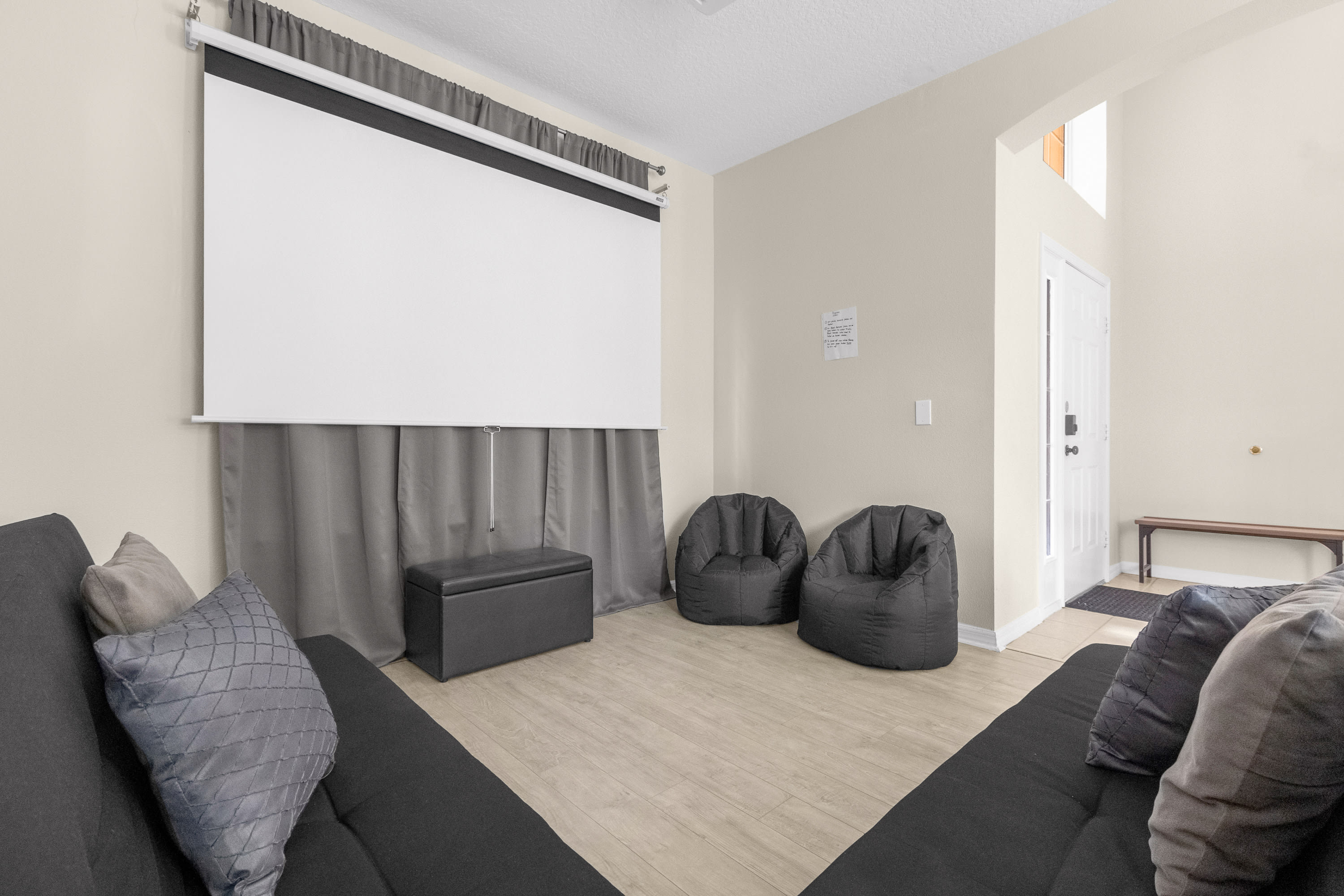 A cozy media room features a large pull-down projector screen, two dark gray bean bag chairs, and two dark gray sofas arranged for comfortable viewing. A black storage ottoman sits beneath the screen, and light wood-look flooring completes the space.