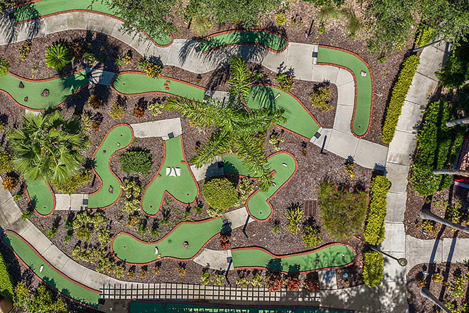 An aerial view of a winding miniature golf course with green turf fairways and concrete paths, surrounded by various plants and palm trees, indicating a recreational amenity.
