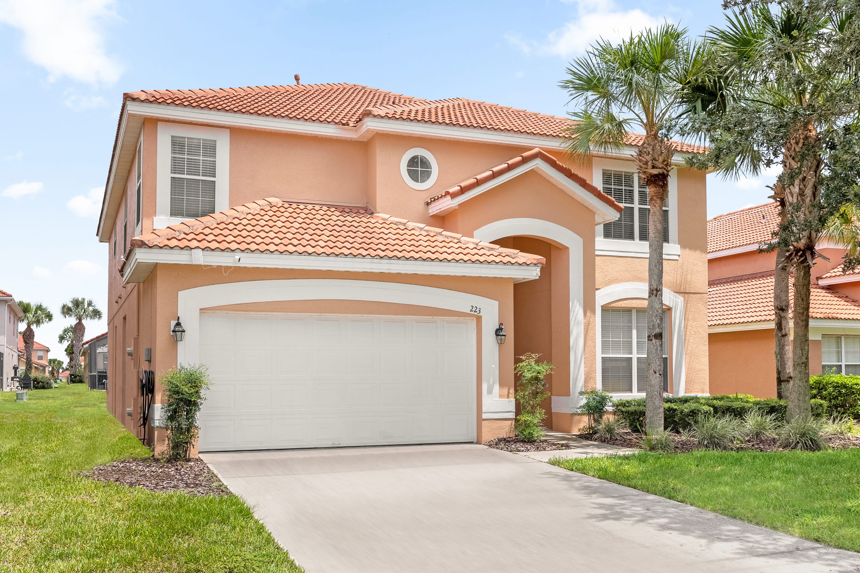 The front exterior of a two-story peach-colored house with a red tile roof features a two-car garage and a paved driveway. Lush green grass and palm trees frame the entrance, with a small porch leading to the front door.