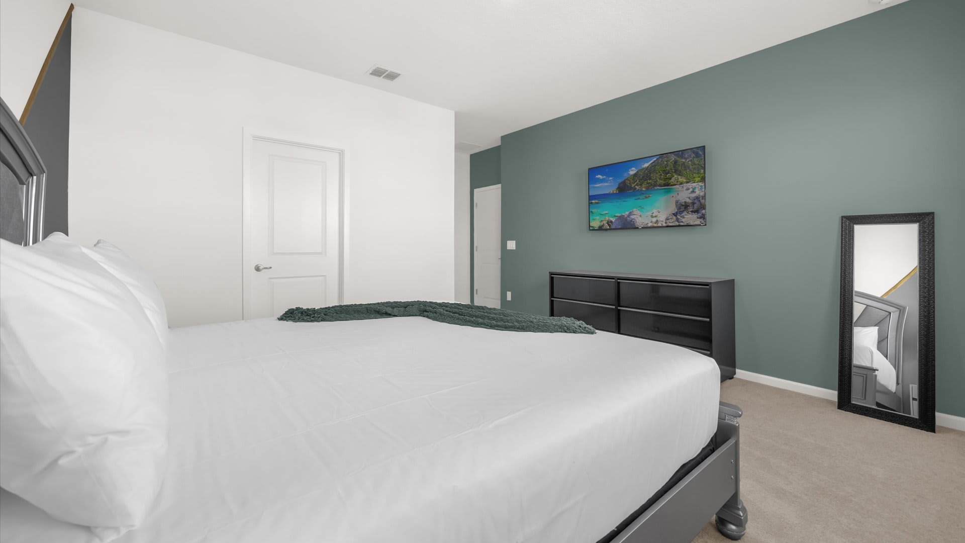 A view of a bedroom with a gray upholstered bed and white bedding, featuring a dark green accent wall with a wall-mounted TV and a black dresser. A full-length mirror leans against the wall, and a white door is visible.