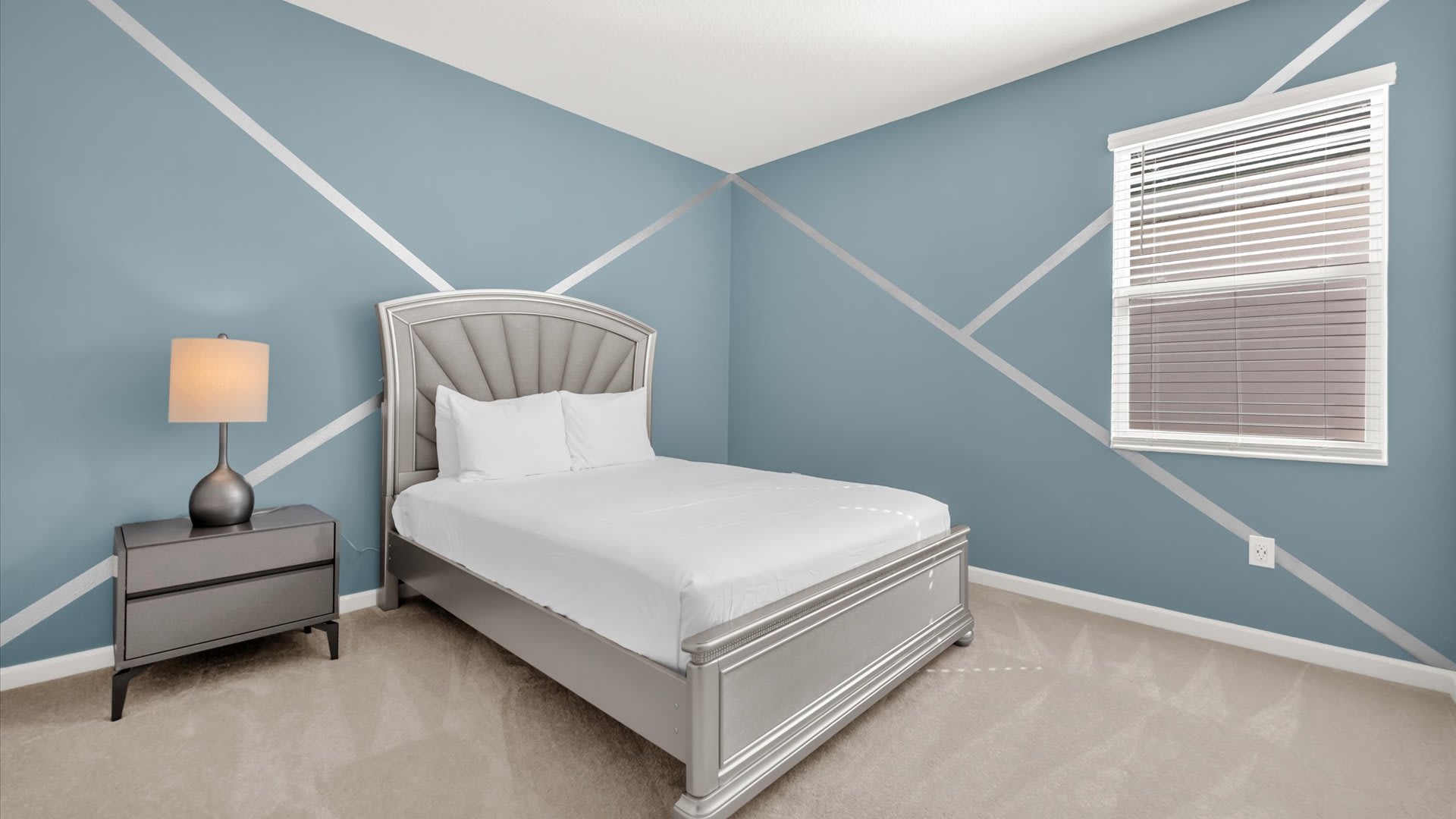 A modern bedroom with a blue accent wall featuring silver geometric stripes, a gray upholstered bed, and a matching gray nightstand with a lamp. A window with blinds offers natural light.