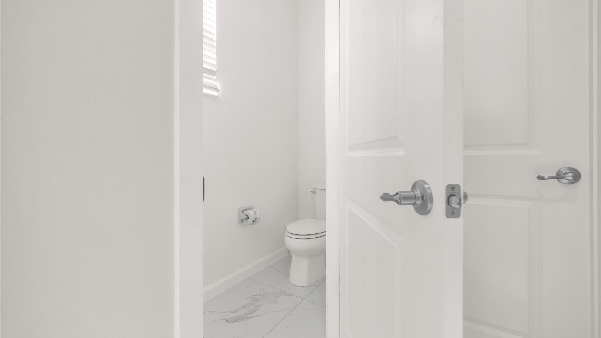 Minimalist powder room with crisp white walls and marble-look tiled floors, featuring a white toilet and a small window providing natural light.