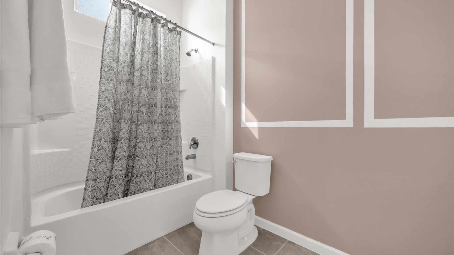 A bathroom with a shower/tub combo featuring a patterned gray shower curtain and a toilet. The wall is painted a light brown, complementing the adjacent bedroom's decor.