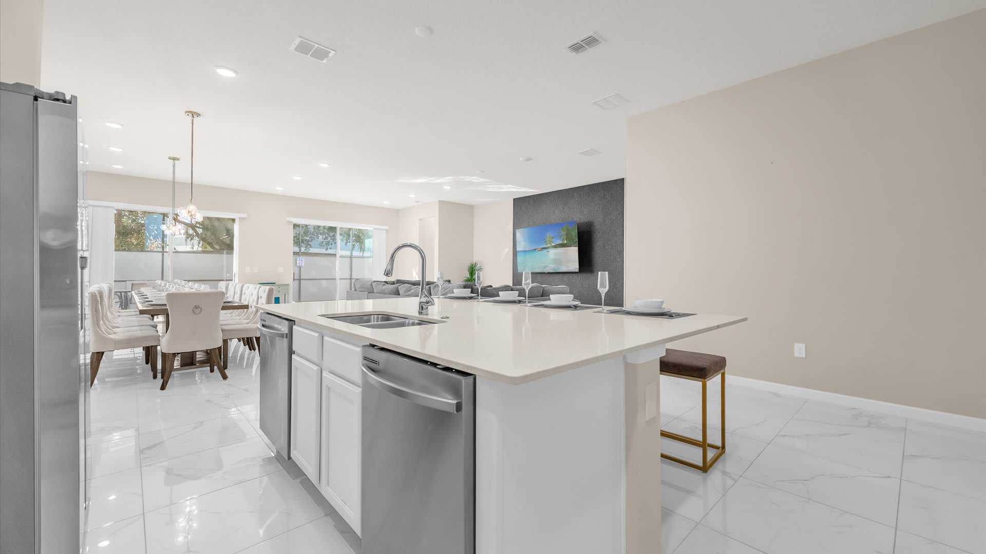 Wide view of the open-concept kitchen island with a sink and dishwasher, looking towards the dining area and living room, all featuring white marble-look tile floors.