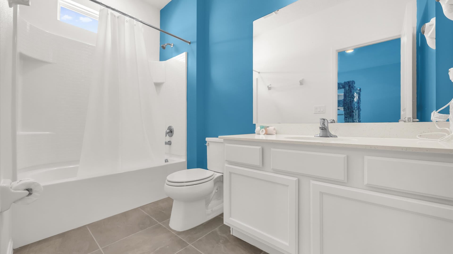 Bright bathroom with a white single vanity, a large mirror, and a white bathtub with a shower curtain, accented by a bold blue wall and light tile flooring.