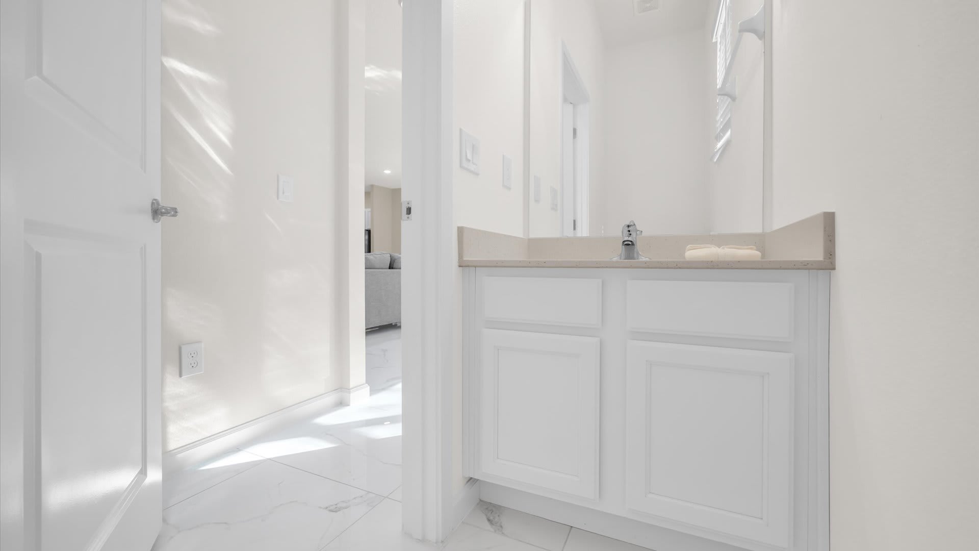 Clean, white hallway leading to a powder room area with a single vanity, light-colored countertop, and white cabinetry, featuring a small mirror.
