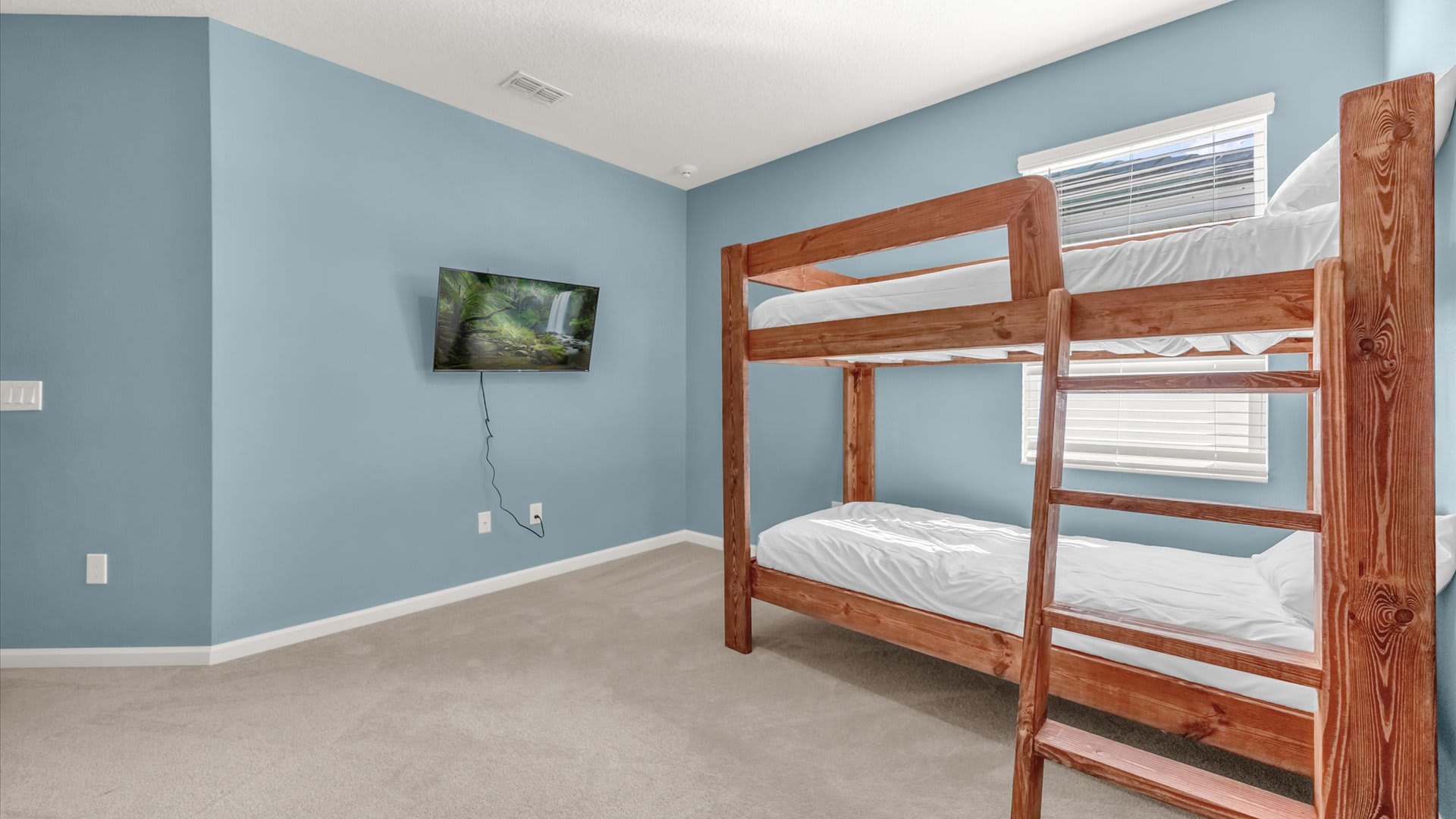 Simple and bright bedroom with light blue walls, a sturdy wooden bunk bed with white bedding, and a wall-mounted television, illuminated by a window with blinds.