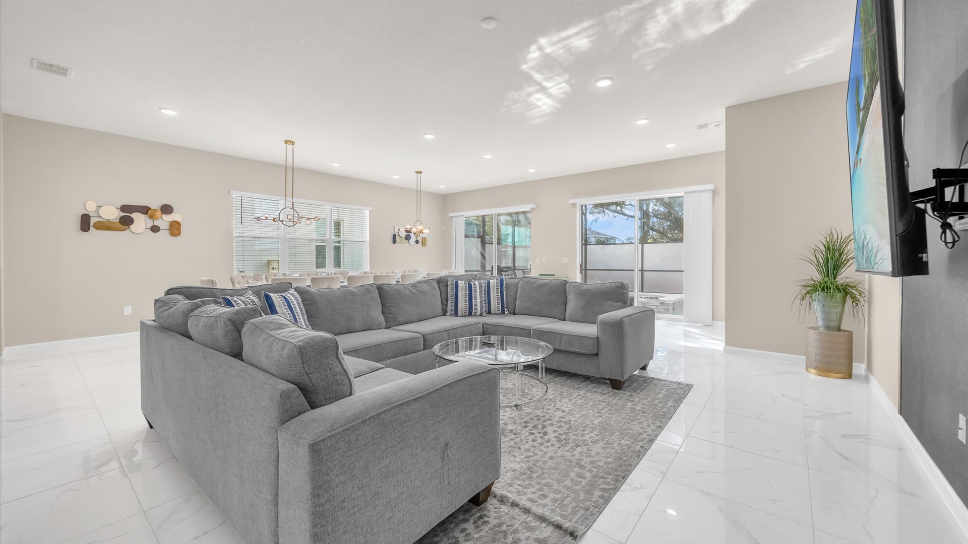 Bright, open-concept living room with a large gray sectional sofa, glass coffee table, and white marble-look tile floors, leading to a dining area and outdoor view.
