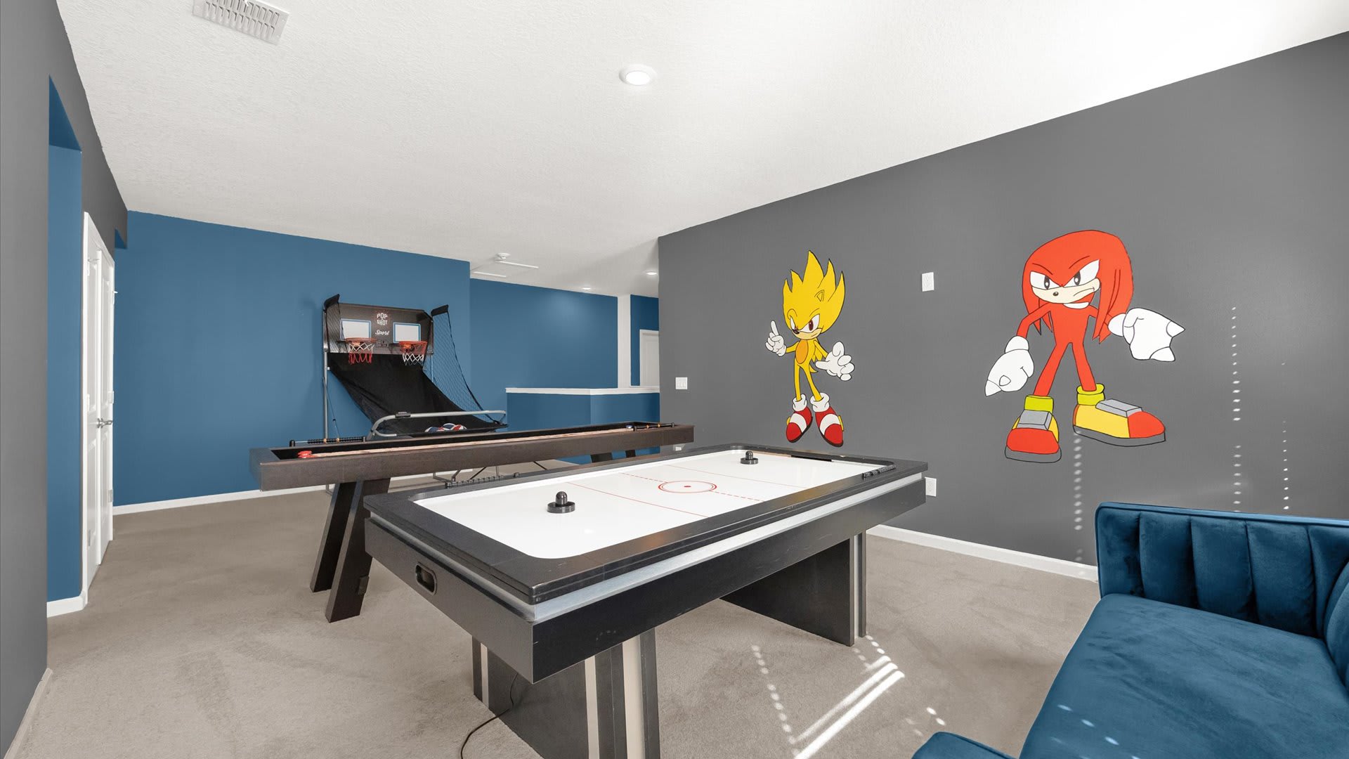 A vibrant game room features a dark blue accent wall with large cartoon character decals (Sonic the Hedgehog and Knuckles), alongside a gray carpeted floor. The room is equipped with an air hockey table, a pool table, and a basketball arcade game, with a blue velvet sofa in the foreground.