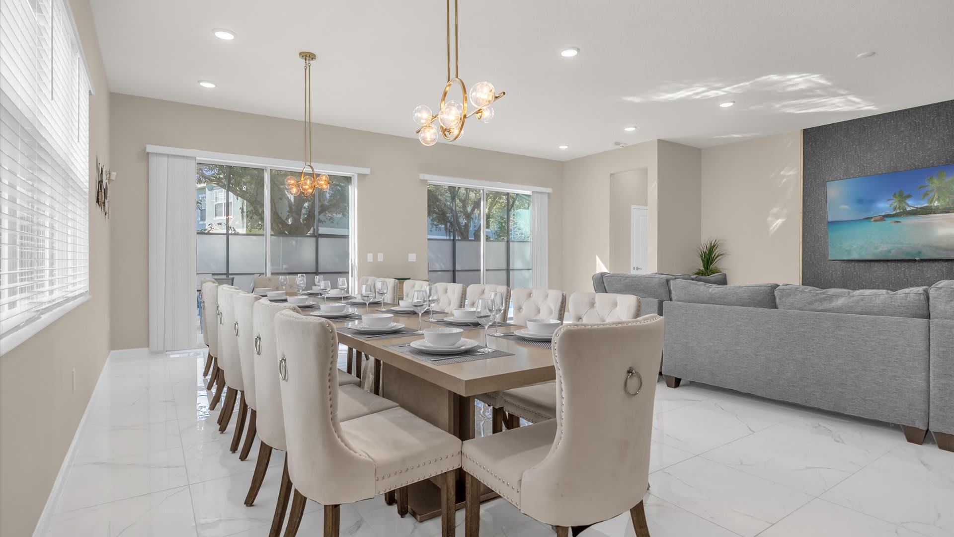 Elegant dining room featuring a long wooden table with 16 upholstered cream chairs, illuminated by two modern gold chandeliers, set on white marble-look tile floors.