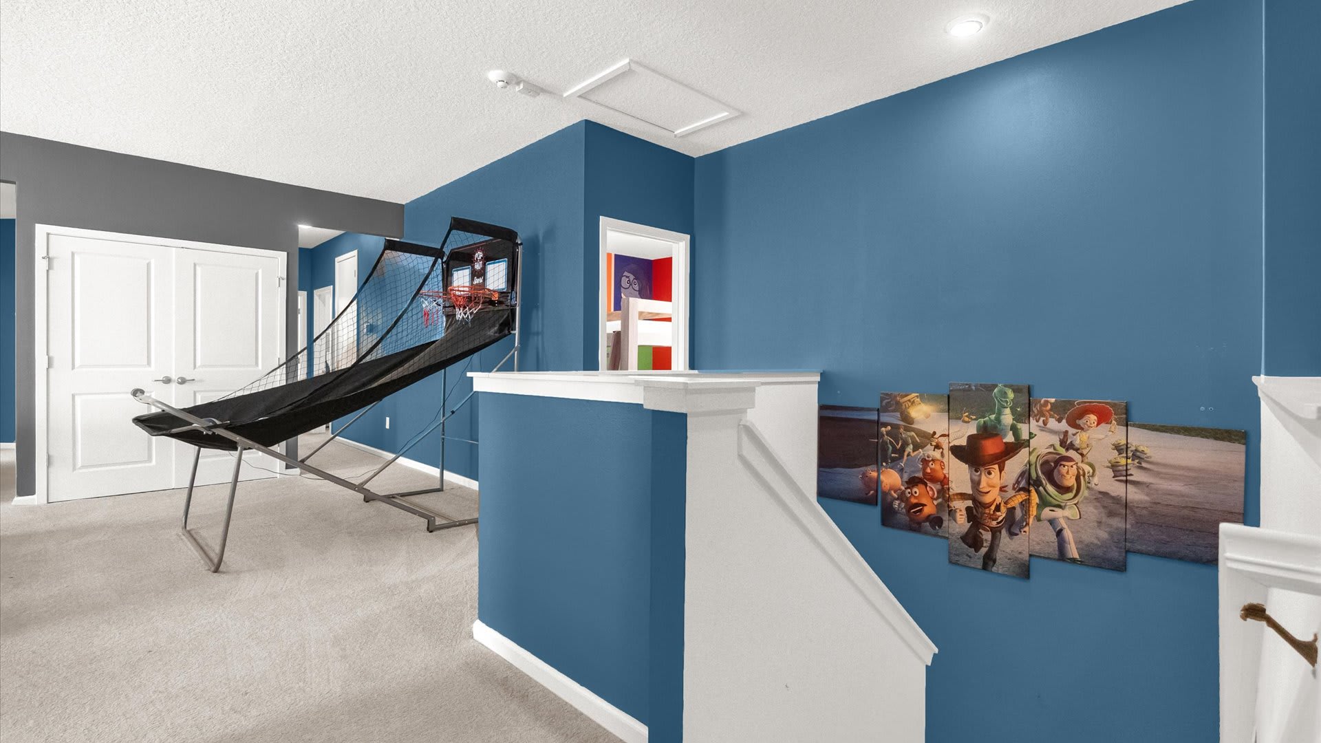 Upper-level landing area with blue walls and carpeted floors, featuring a basketball arcade game and Toy Story wall art, leading to other rooms.