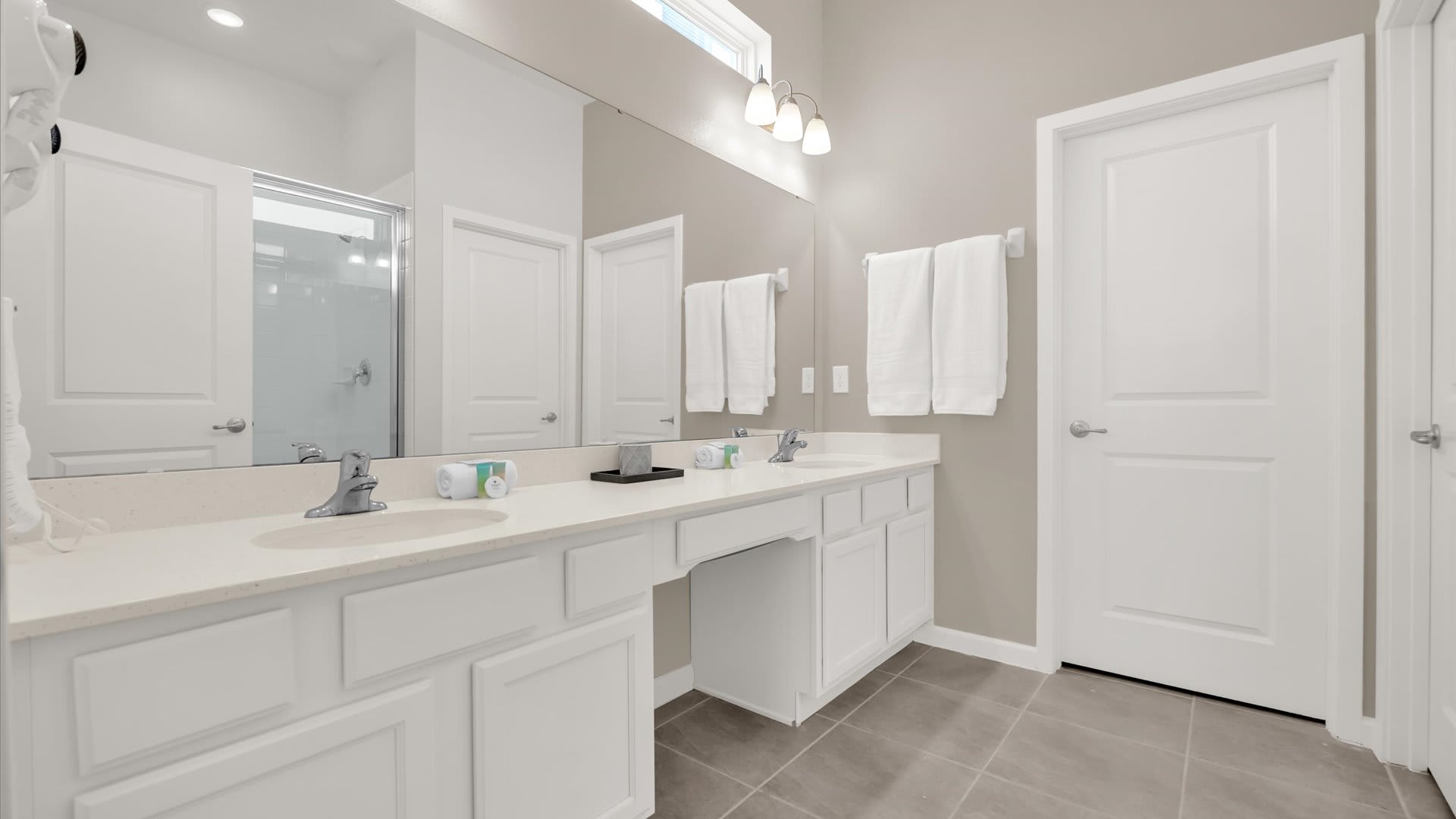 Spacious en-suite bathroom featuring a long double vanity with white cabinets, light countertops, and two sinks. White towels are neatly folded, and a large mirror spans the length of the vanity.