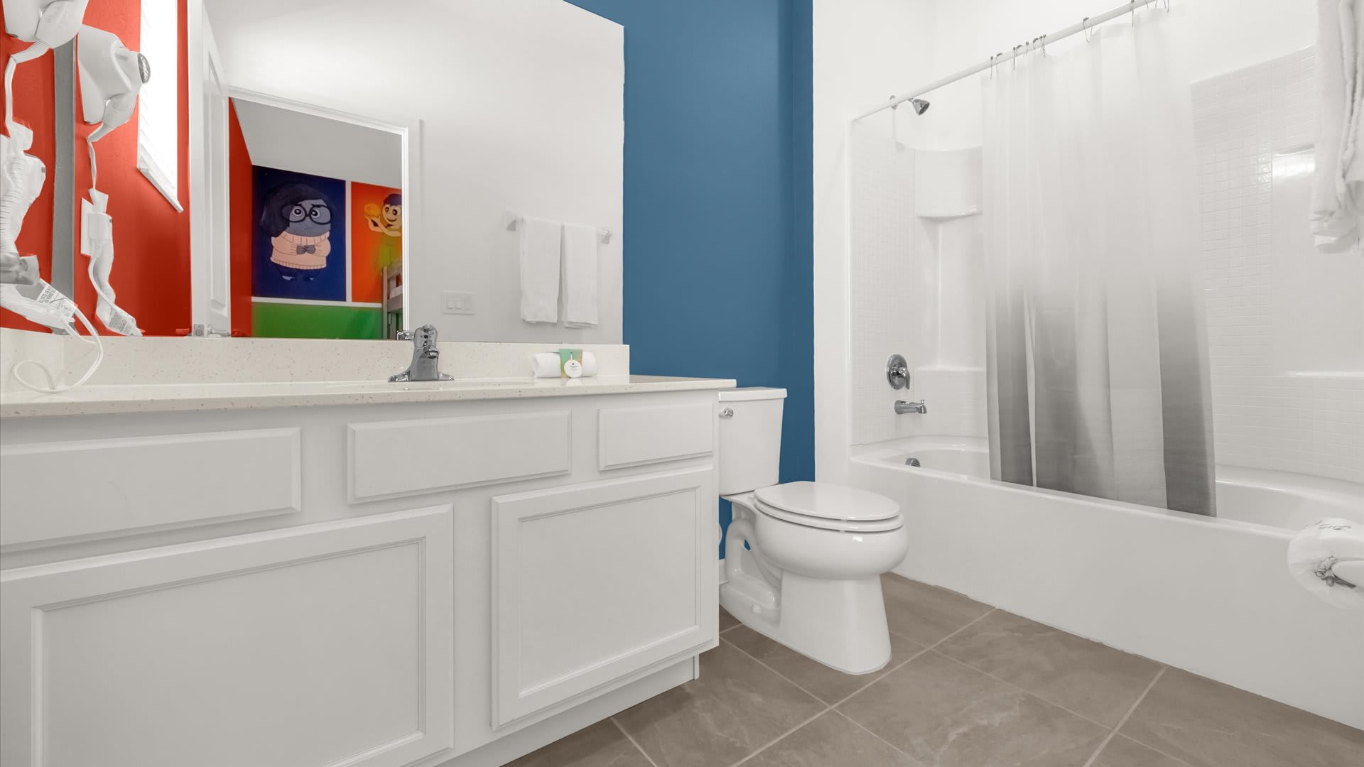 Kids' bathroom with a white single vanity, light countertop, and a toilet. A shower-tub combo with a gray and white curtain is visible, and the mirror reflects the colorful themed bedroom.