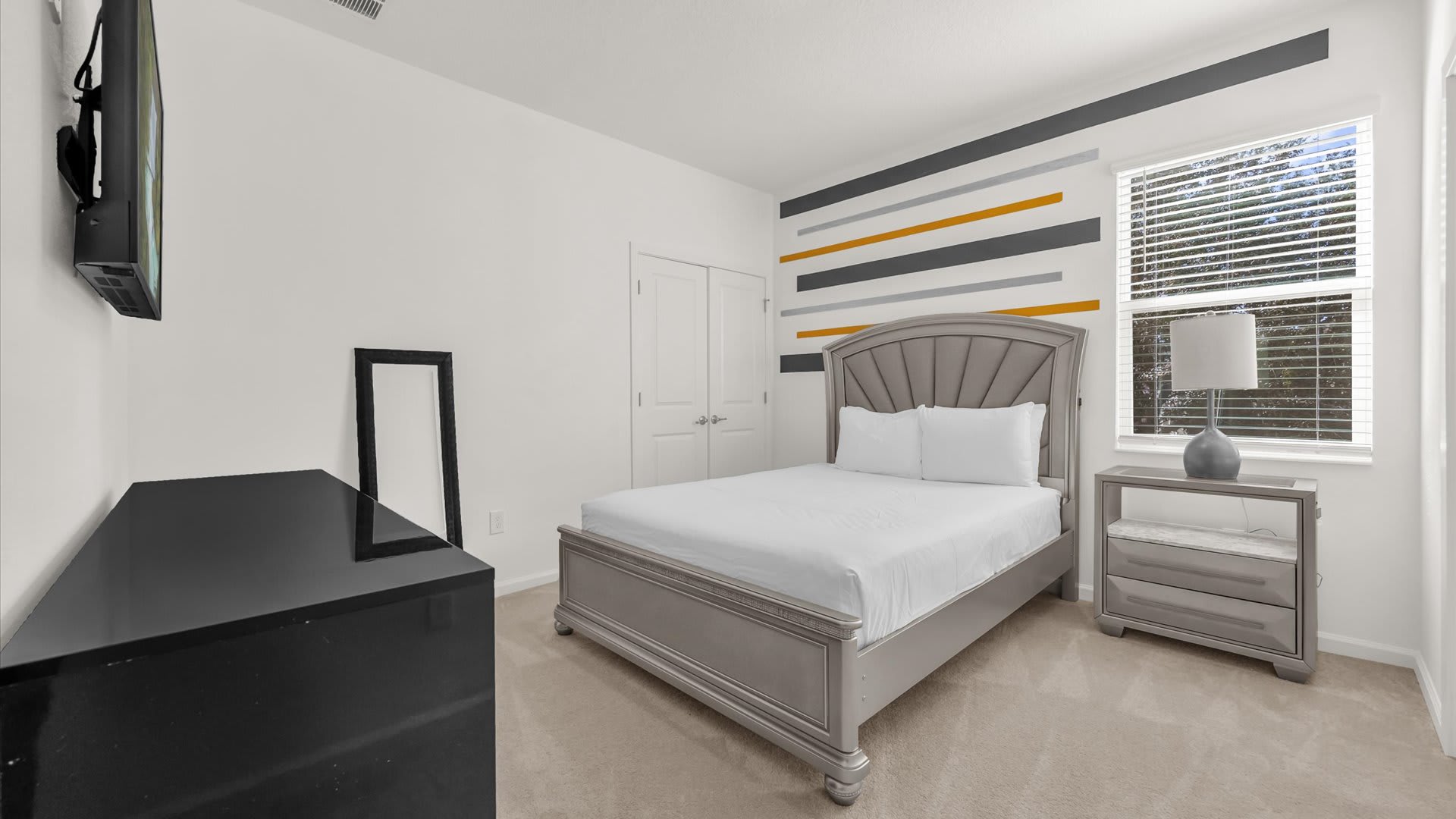 A bright bedroom with white walls, a gray upholstered bed, and a black dresser with a TV mounted above it. A decorative striped accent wall in gray, yellow, and orange adds a modern touch.