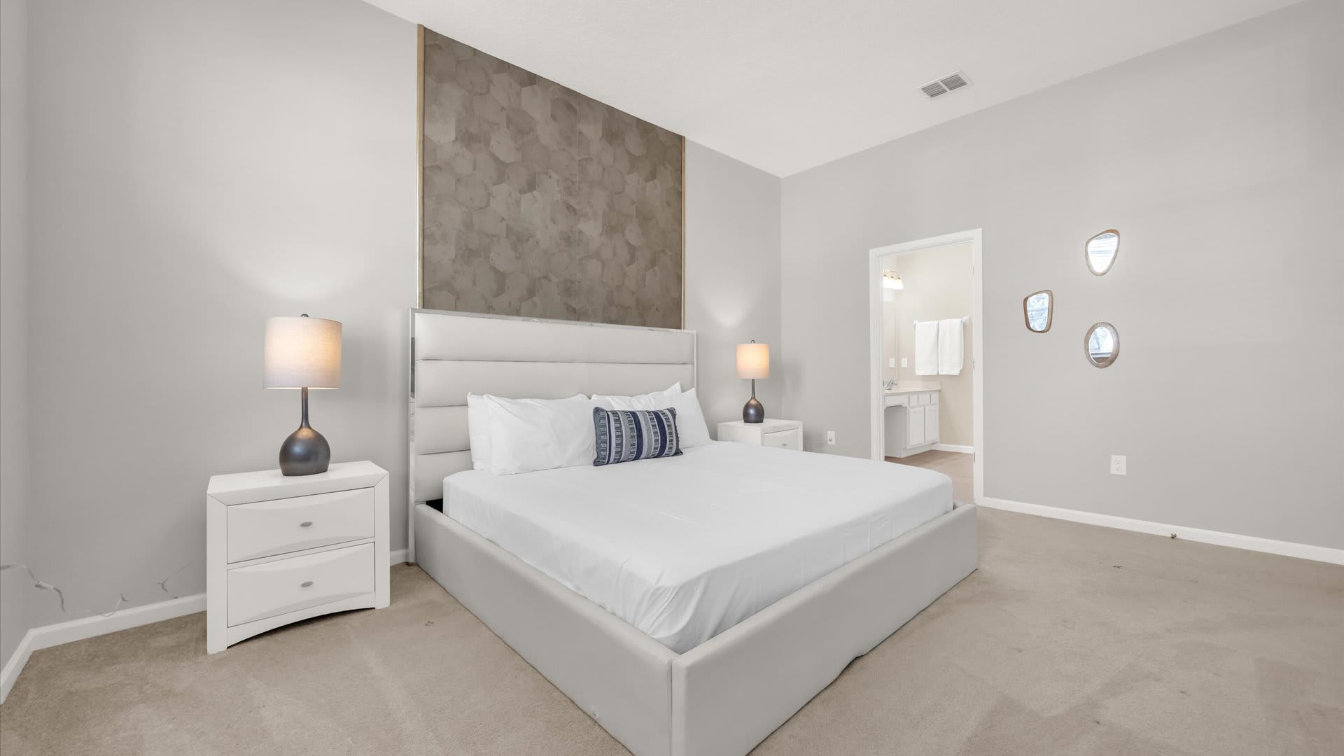 Bright master bedroom featuring a king-sized bed with a white upholstered headboard, two white nightstands with modern lamps, and a textured geometric accent wall behind the bed. A doorway leads to an en-suite bathroom.