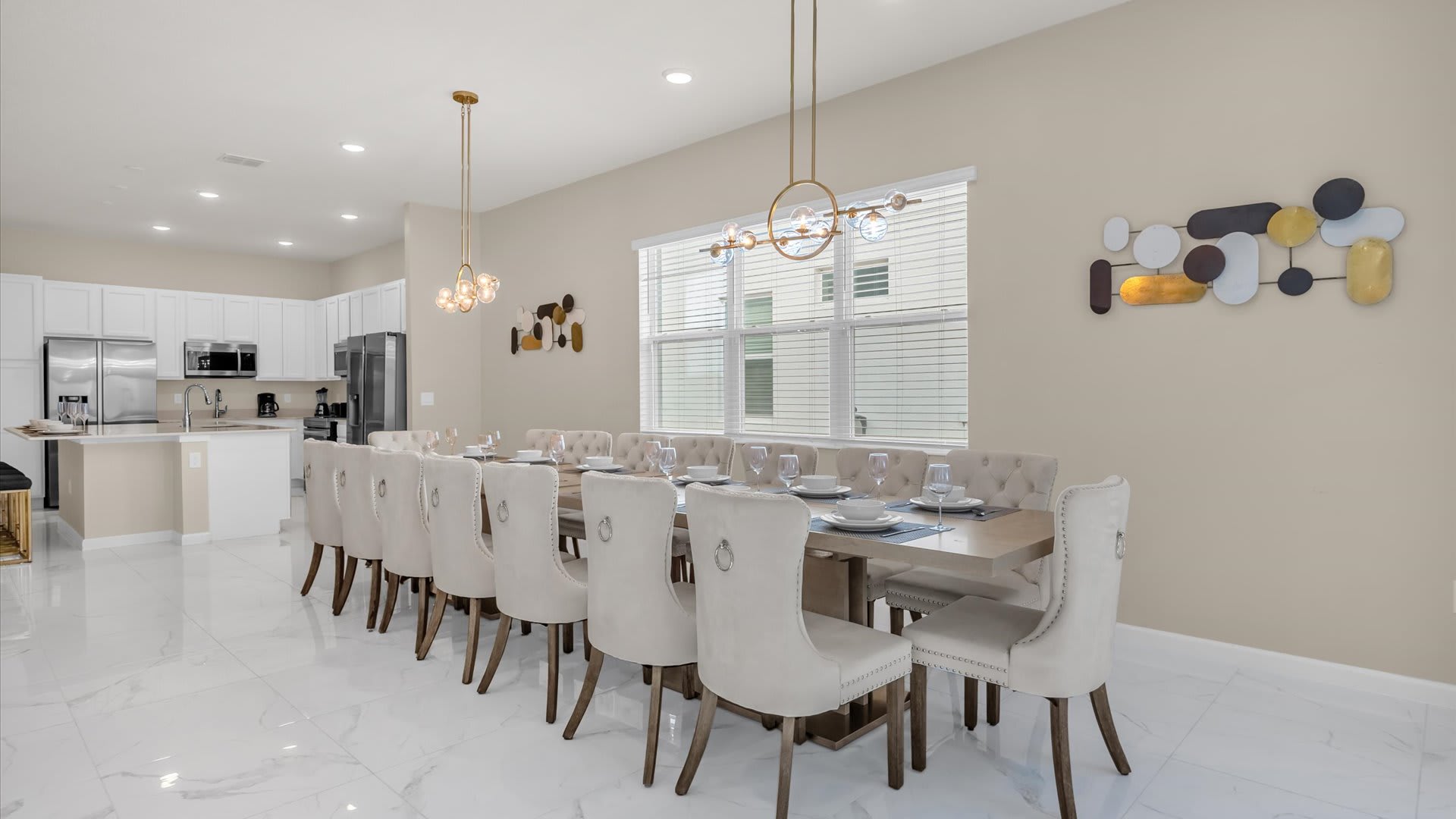 A spacious dining area features a long rectangular wooden table with seating for many, surrounded by upholstered dining chairs. Modern gold chandeliers hang above, and a large window with blinds overlooks the exterior, with a contemporary kitchen visible in the background.
