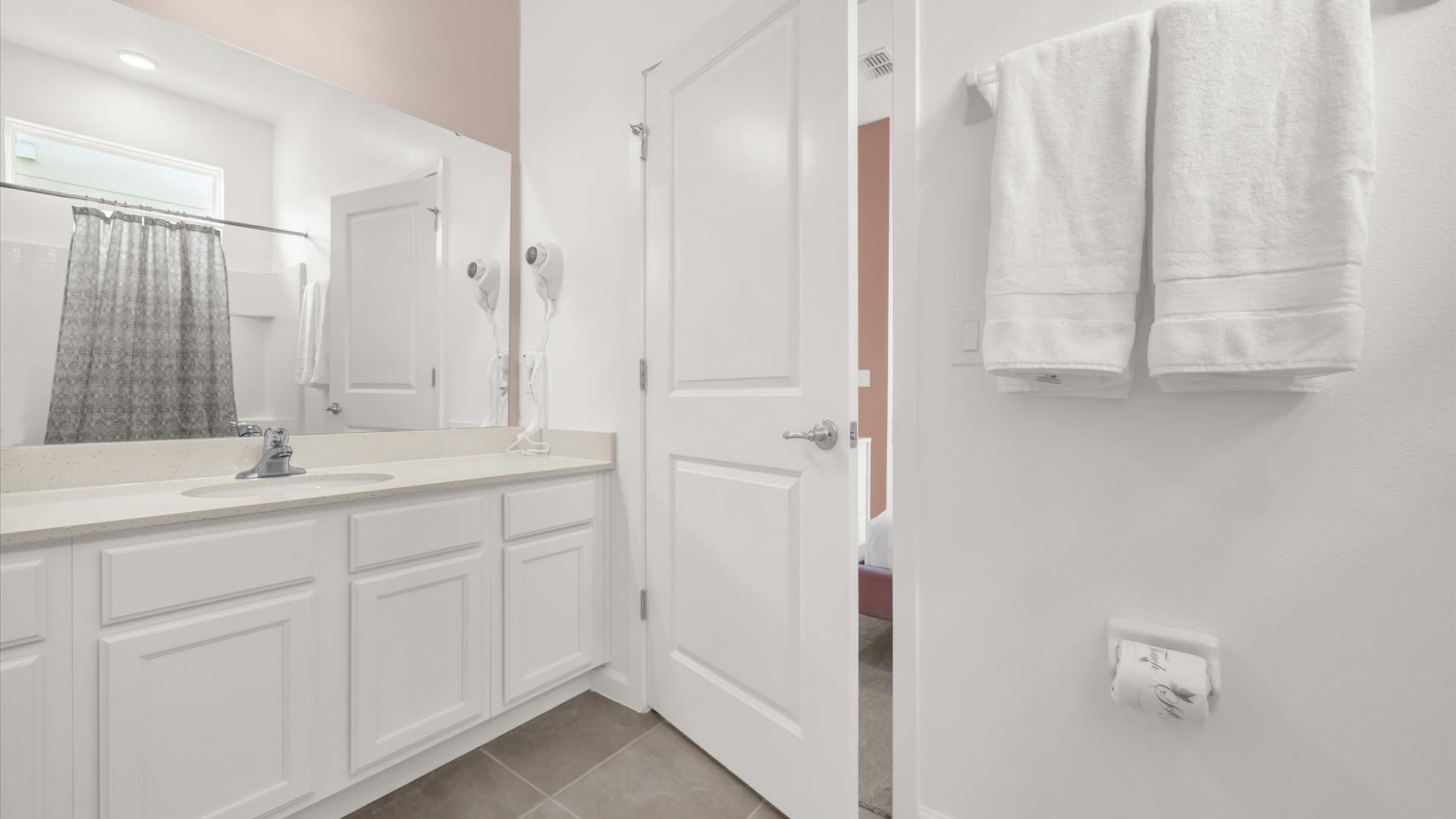 A bright bathroom with a white single vanity and light countertop, a large mirror, and a white door leading to the adjacent bedroom. White towels are neatly hung on the wall.