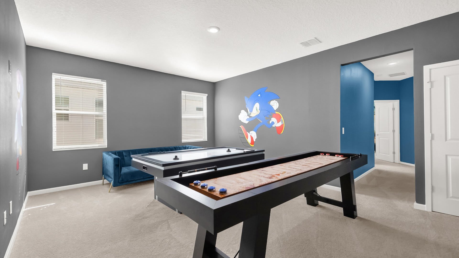 Game room with dark gray walls and carpeted floors, featuring a shuffleboard table, an air hockey table, and a blue velvet sofa, with a Sonic the Hedgehog wall decal.