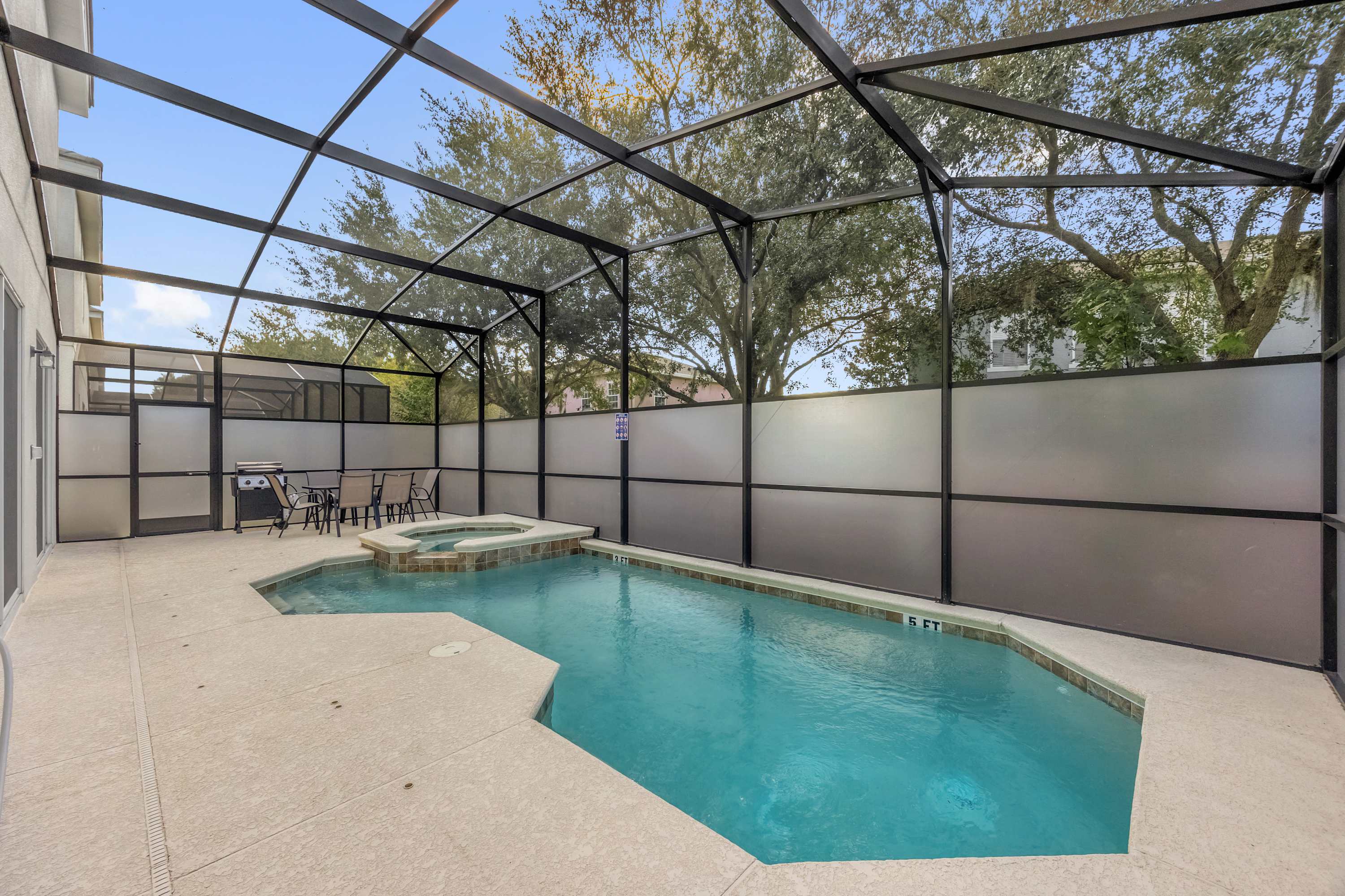 A screened-in private pool area features a rectangular pool with an integrated hot tub, surrounded by a light-colored concrete deck. A small dining table with chairs and a BBQ grill are visible in the corner, all enclosed by a black metal screen structure and frosted privacy panels.