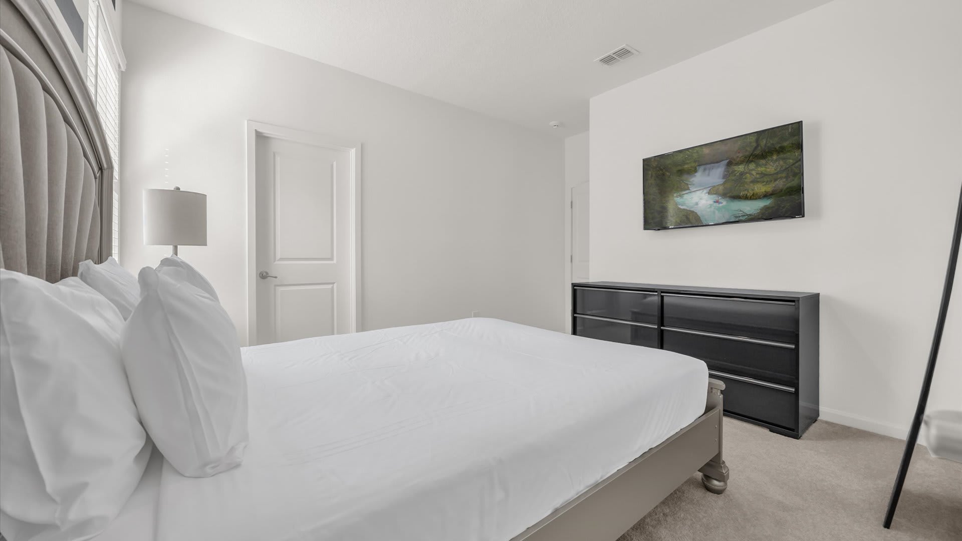 View from the bed in a bright bedroom, showing a wall-mounted TV above a black dresser and a white door. The room features white walls and a light-colored carpet.