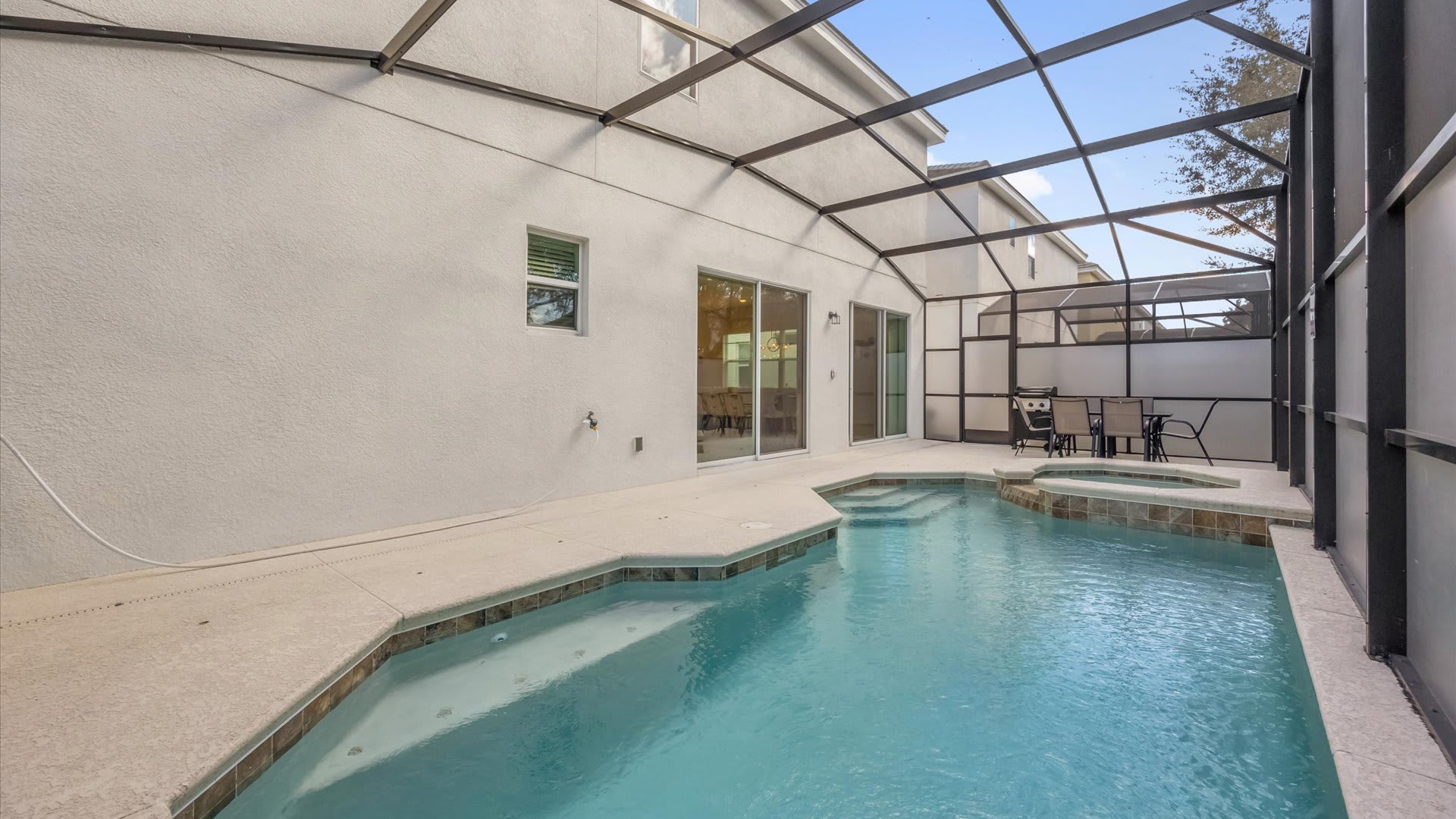 A wide view of the screened-in private pool area reveals the house's exterior with sliding glass doors leading to the pool deck. The rectangular pool and hot tub are central, with ample concrete patio space for lounging, all under the protective screen enclosure.