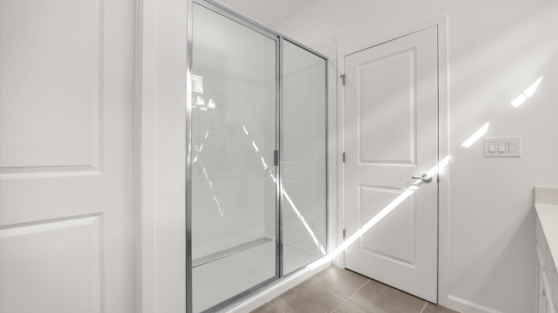 A bright bathroom showcasing a spacious glass-enclosed shower with white subway tiles. White doors are visible on either side of the shower, and sunlight streams through a window.
