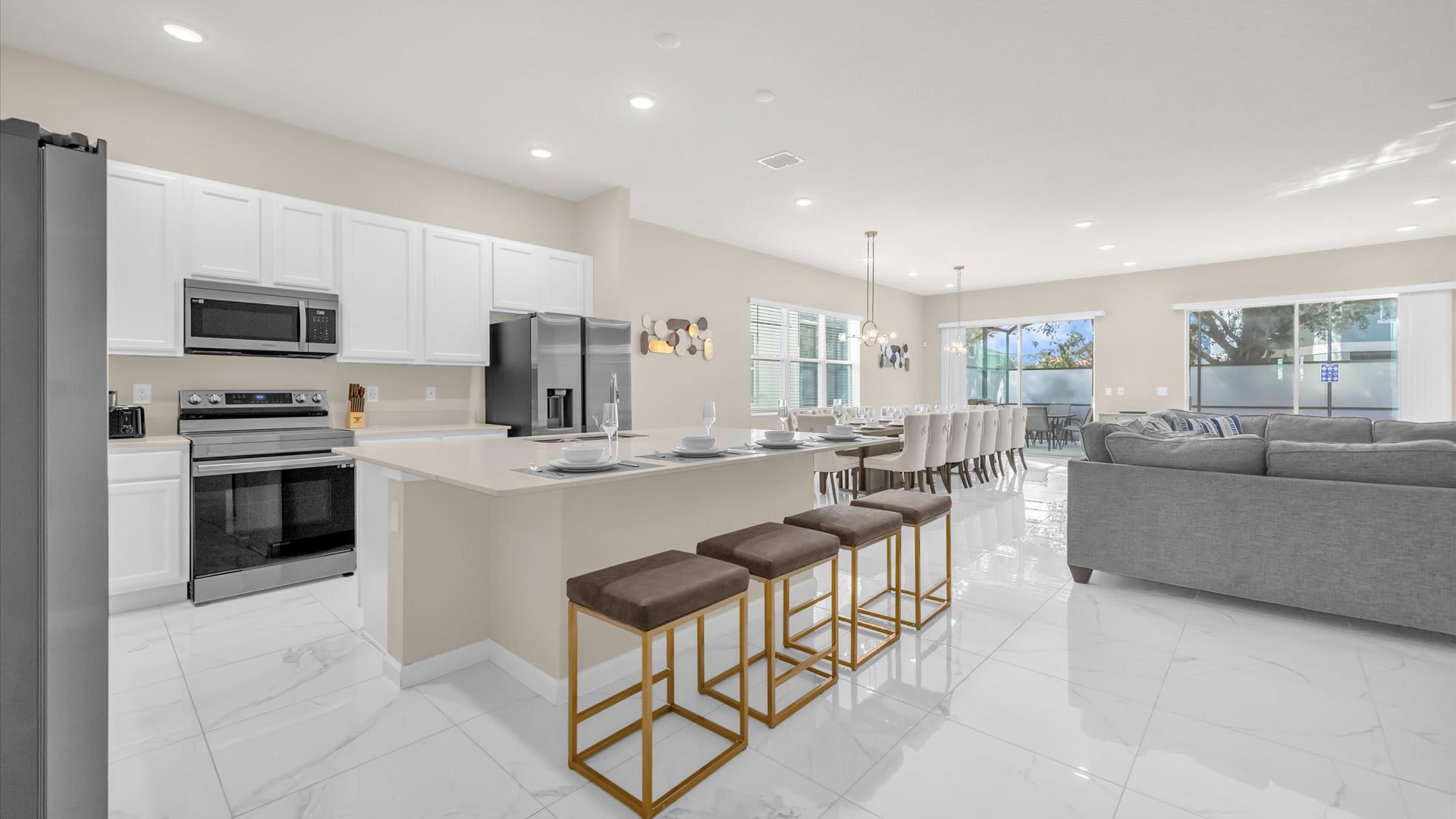 Open-concept kitchen with a large island and bar seating, connecting to the dining area and living room, featuring white cabinets, stainless steel appliances, and marble-look tile floors.