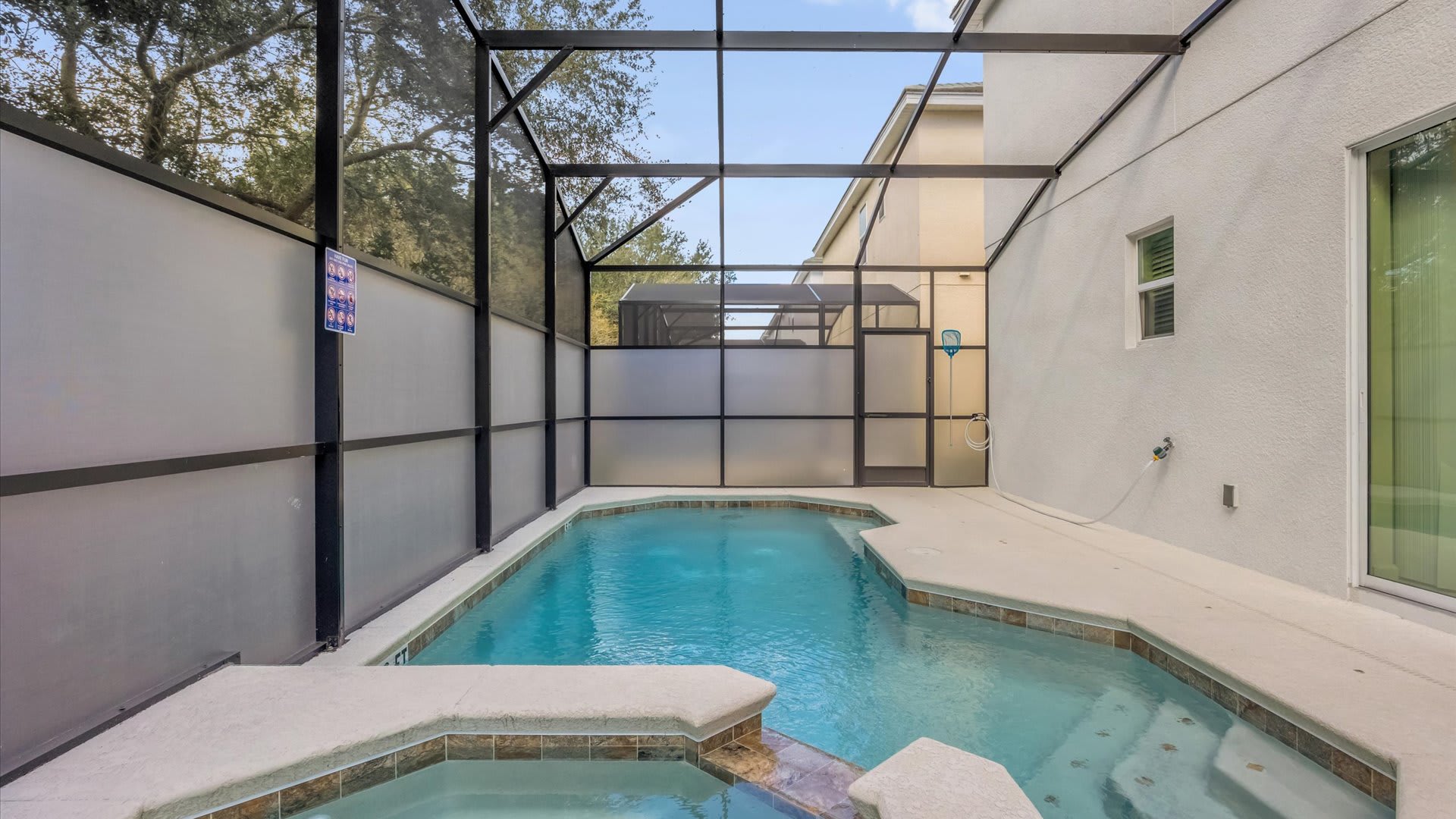 A screened-in private pool area shows a rectangular pool with an integrated hot tub, set against the side of the house. The enclosure features frosted privacy panels and a gate, with a small basketball hoop visible in the corner, offering a compact recreational space.
