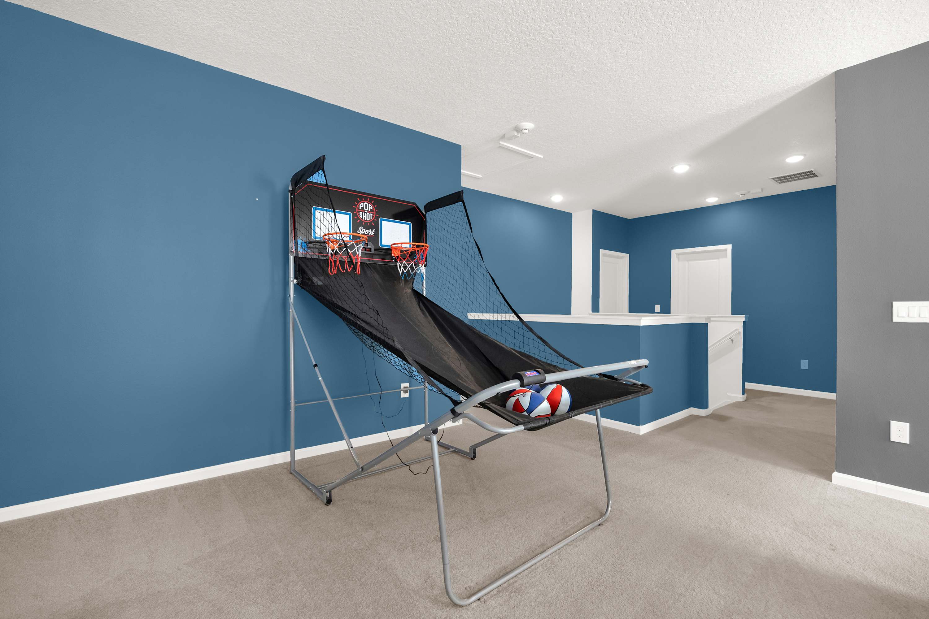 Upper-level game room with blue walls and carpeted floors, featuring a dual basketball arcade game and a view of the staircase landing.