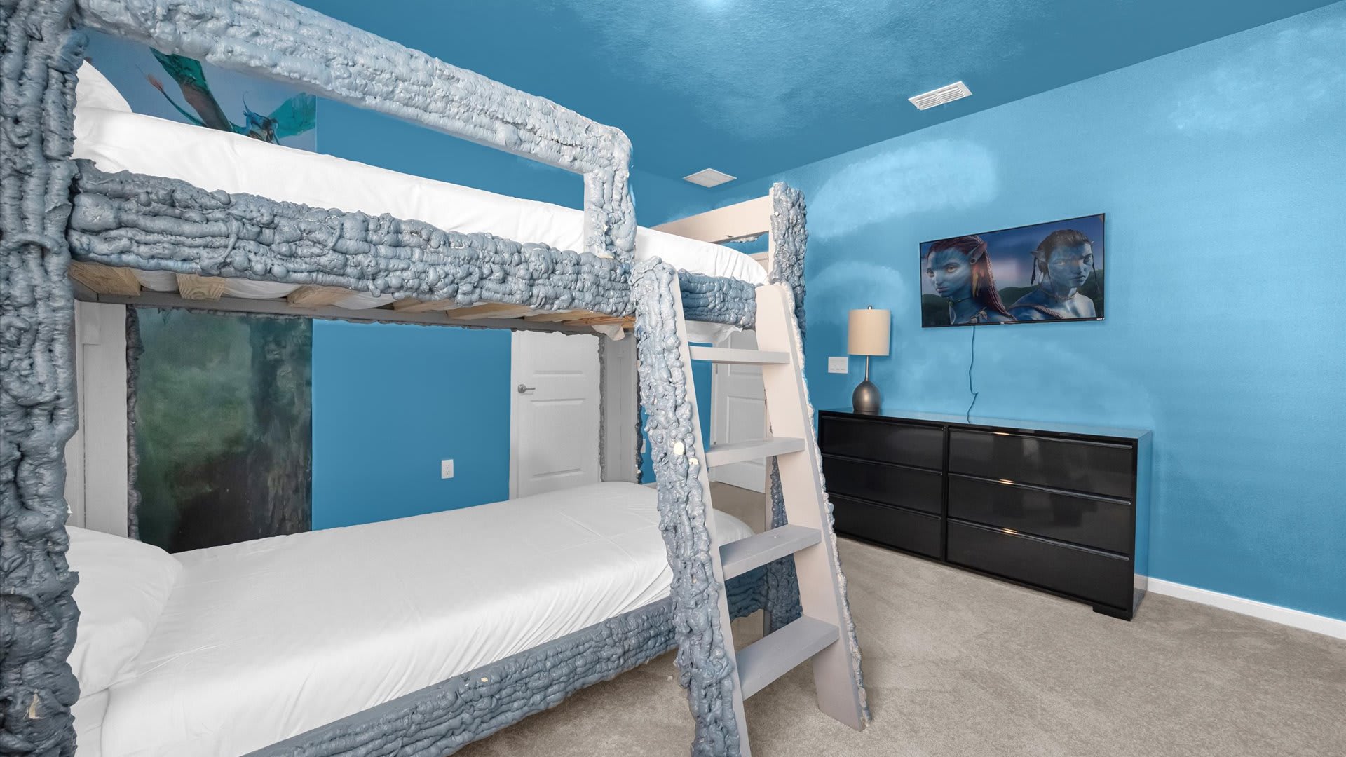 View of the Avatar-themed bedroom with a blue textured bunk bed and a black dresser topped with a TV. The walls and ceiling are painted in shades of blue, enhancing the immersive theme.