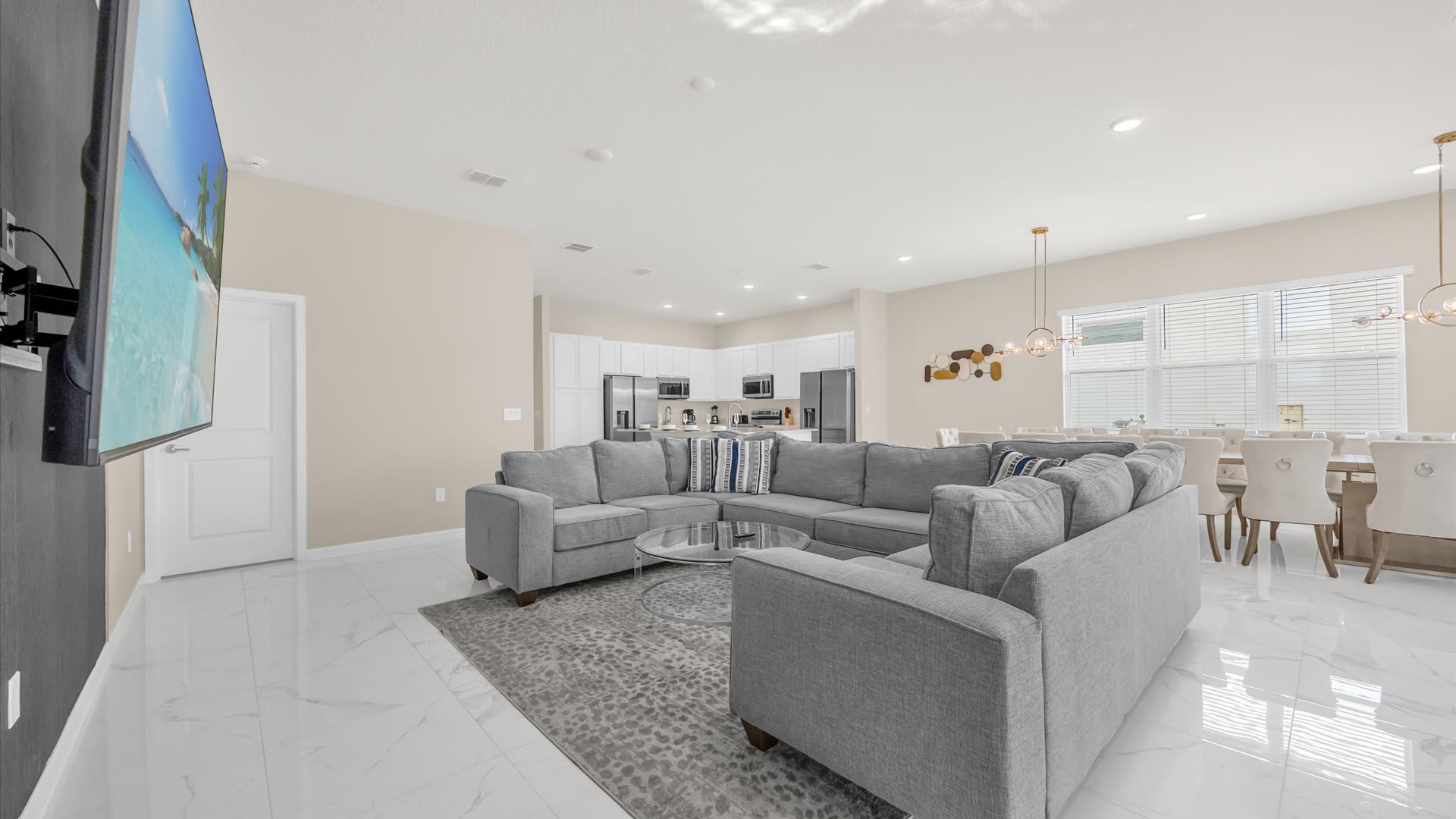 A bright open-concept living room features a large gray sectional sofa arranged around a glass coffee table on a patterned rug. A wall-mounted TV is visible on a dark accent wall, with the dining area and modern kitchen in the background, all set on a glossy white tile floor.