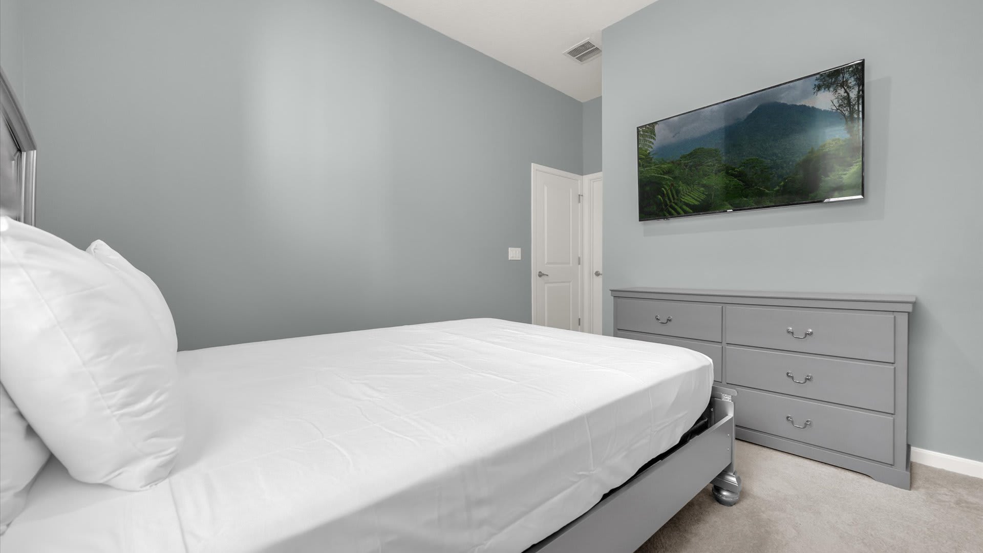 View from the bed in a light gray bedroom, showing a wall-mounted TV above a gray dresser and a white door. The room maintains a calm, neutral color palette.