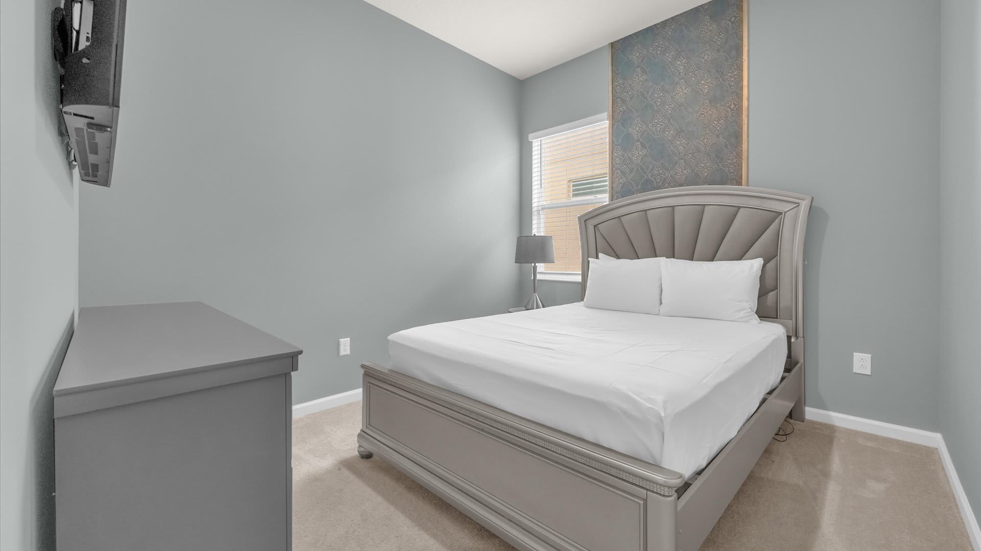A serene bedroom featuring light gray walls, a gray upholstered bed with white bedding, and a matching gray dresser. A window with blinds and a decorative wall panel add visual interest.