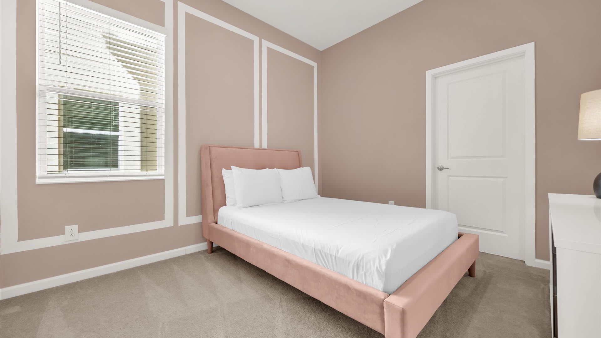 A cozy bedroom featuring a pink upholstered bed with white bedding, set against a light brown accent wall with white decorative trim. A window with blinds and a white door are also visible.