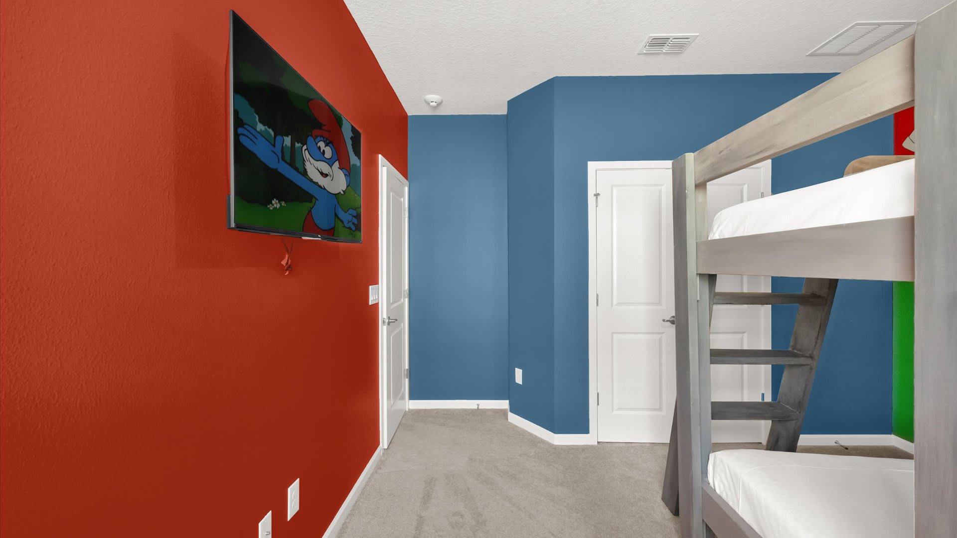 Another angle of the colorful kids' bedroom, showcasing a wall-mounted TV on a red wall featuring a Smurf character. A wooden bunk bed is visible on the right.