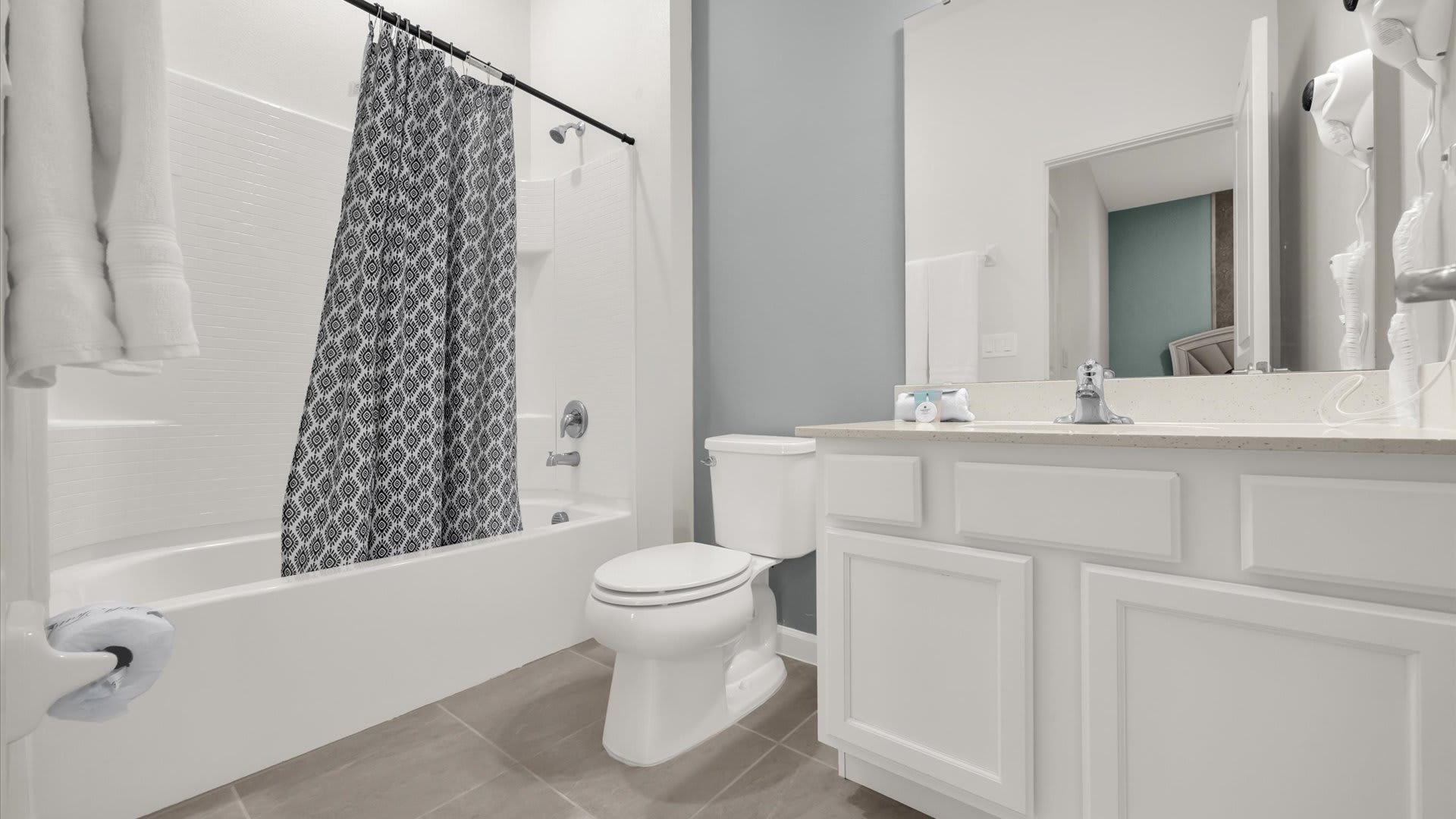 A bright bathroom with a white single vanity, a toilet, and a shower/tub combo with a patterned gray shower curtain. A light blue accent wall behind the toilet adds a subtle pop of color.