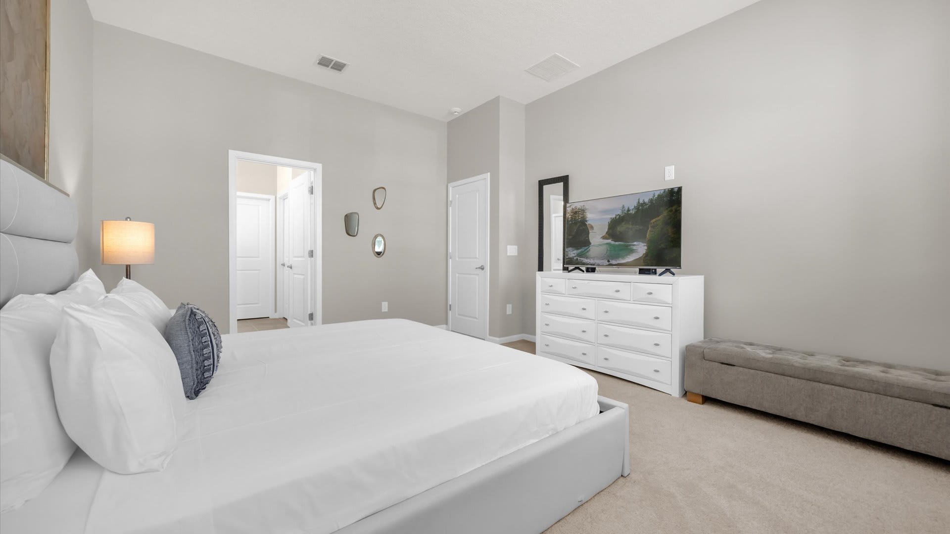 Perspective of the master bedroom showing the foot of the king bed, a gray upholstered bench, and a white dresser with a large TV. Light gray walls and carpeted flooring create a serene atmosphere.