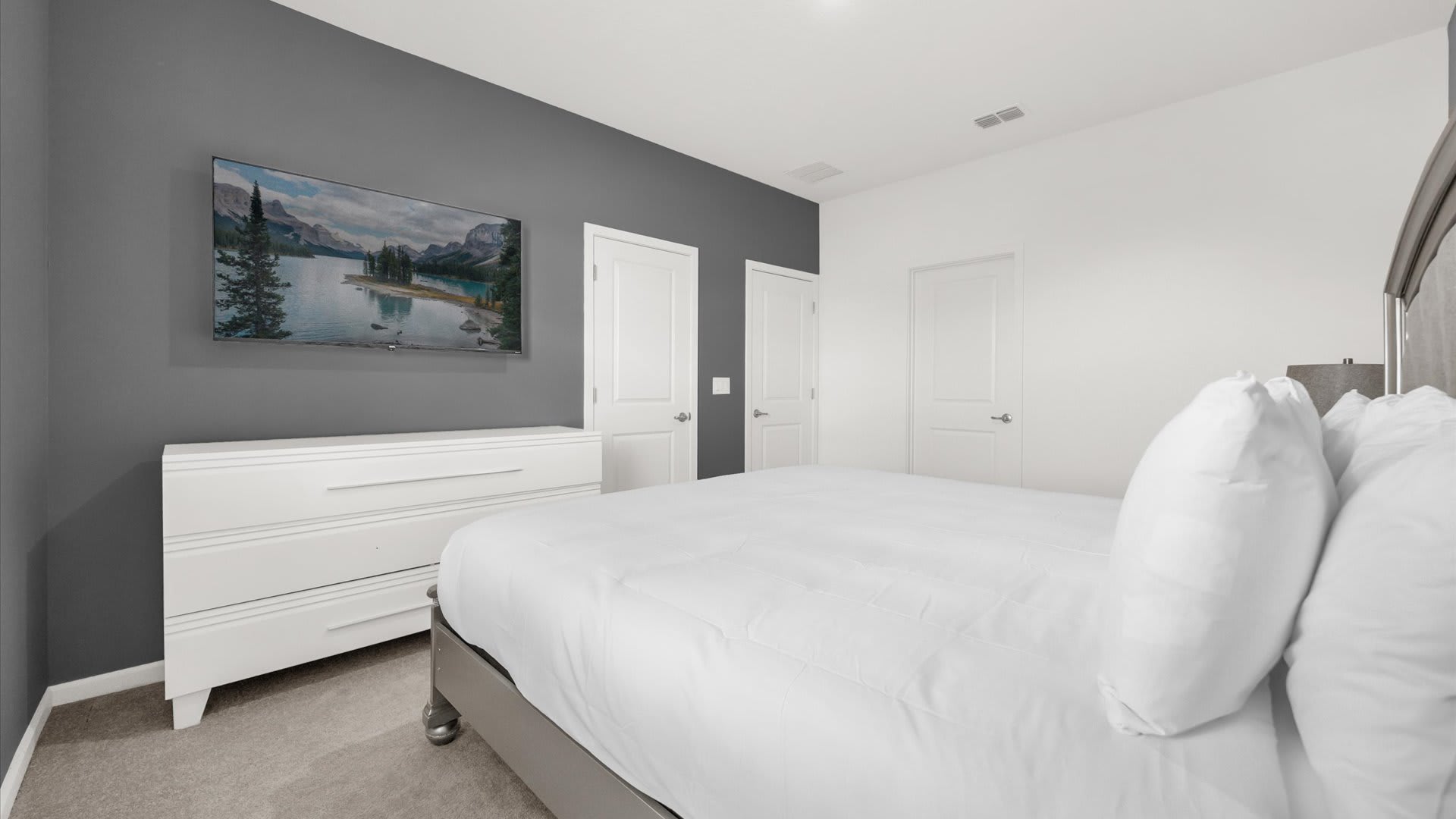 A modern bedroom featuring a queen-sized bed with white bedding, a white dresser, and a wall-mounted TV against a dark gray accent wall. The room has light gray carpet and white doors.