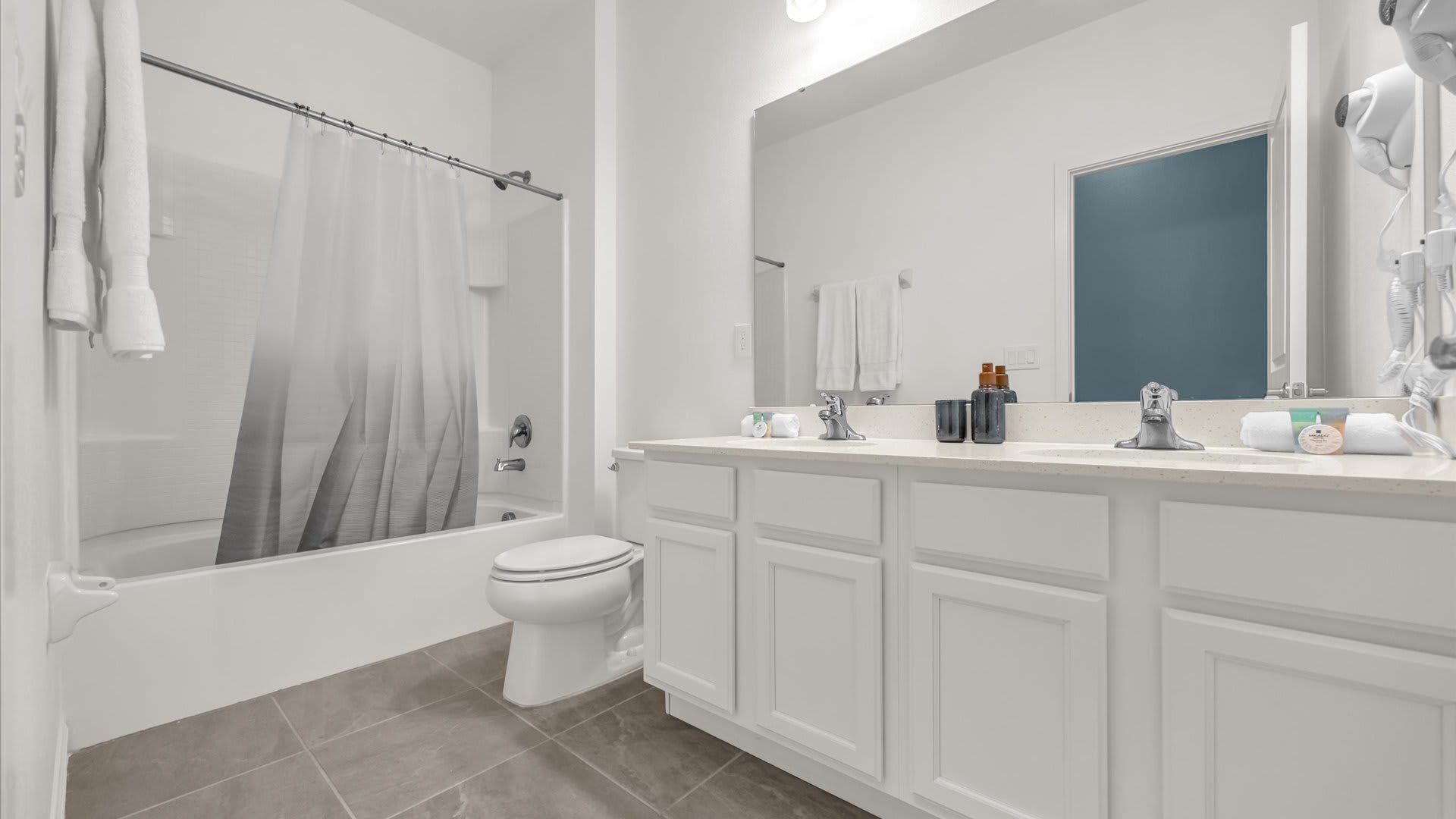 Modern bathroom featuring a double white vanity with two sinks, a large mirror, and a white bathtub with a shower curtain, set against light gray tiled flooring.