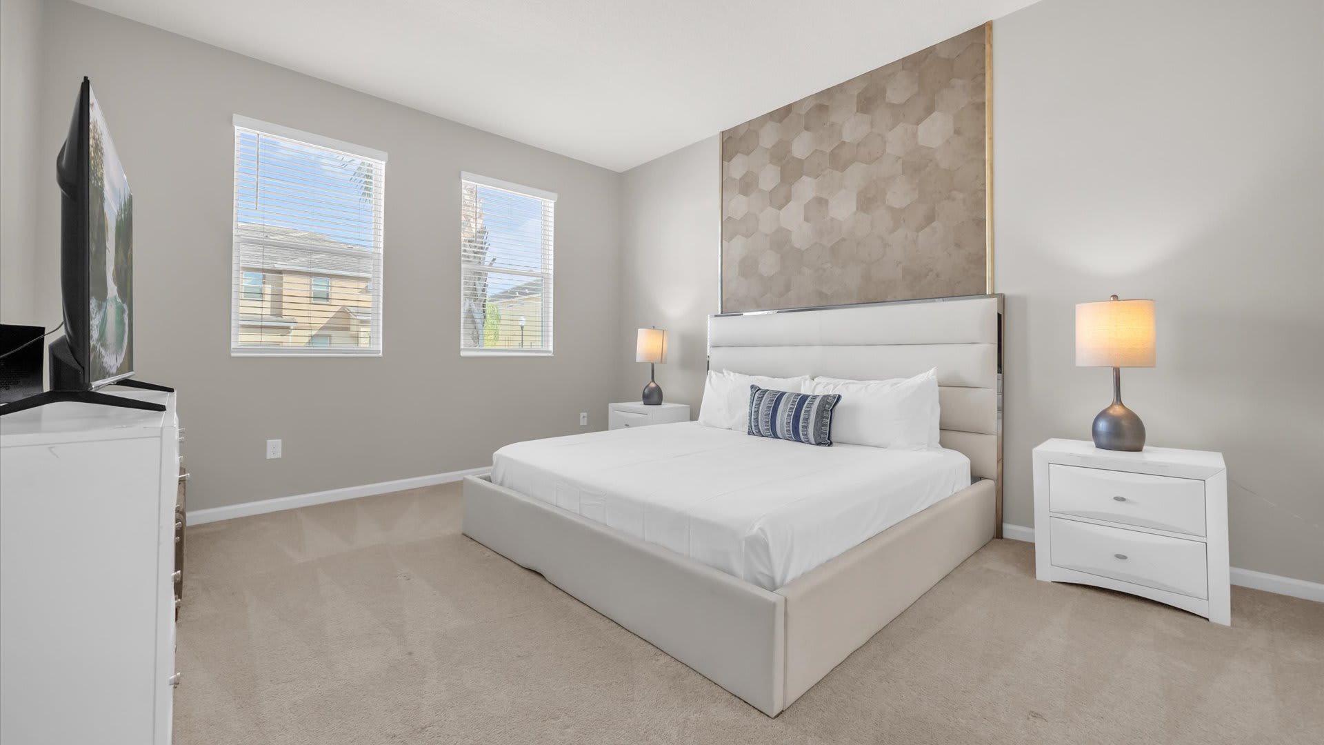 View of the master bedroom with a king-sized bed, white dresser with a TV, and two windows with blinds. The room has light gray walls and a textured geometric accent wall behind the bed.