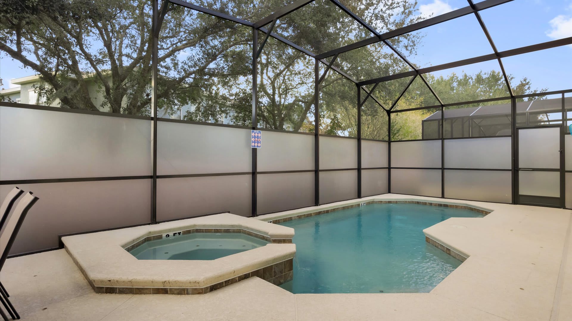 A private screened-in pool area features a rectangular pool with an attached hot tub, surrounded by a light-colored concrete deck. The enclosure has frosted privacy panels along the bottom, with trees visible through the upper screen, creating a secluded outdoor space.