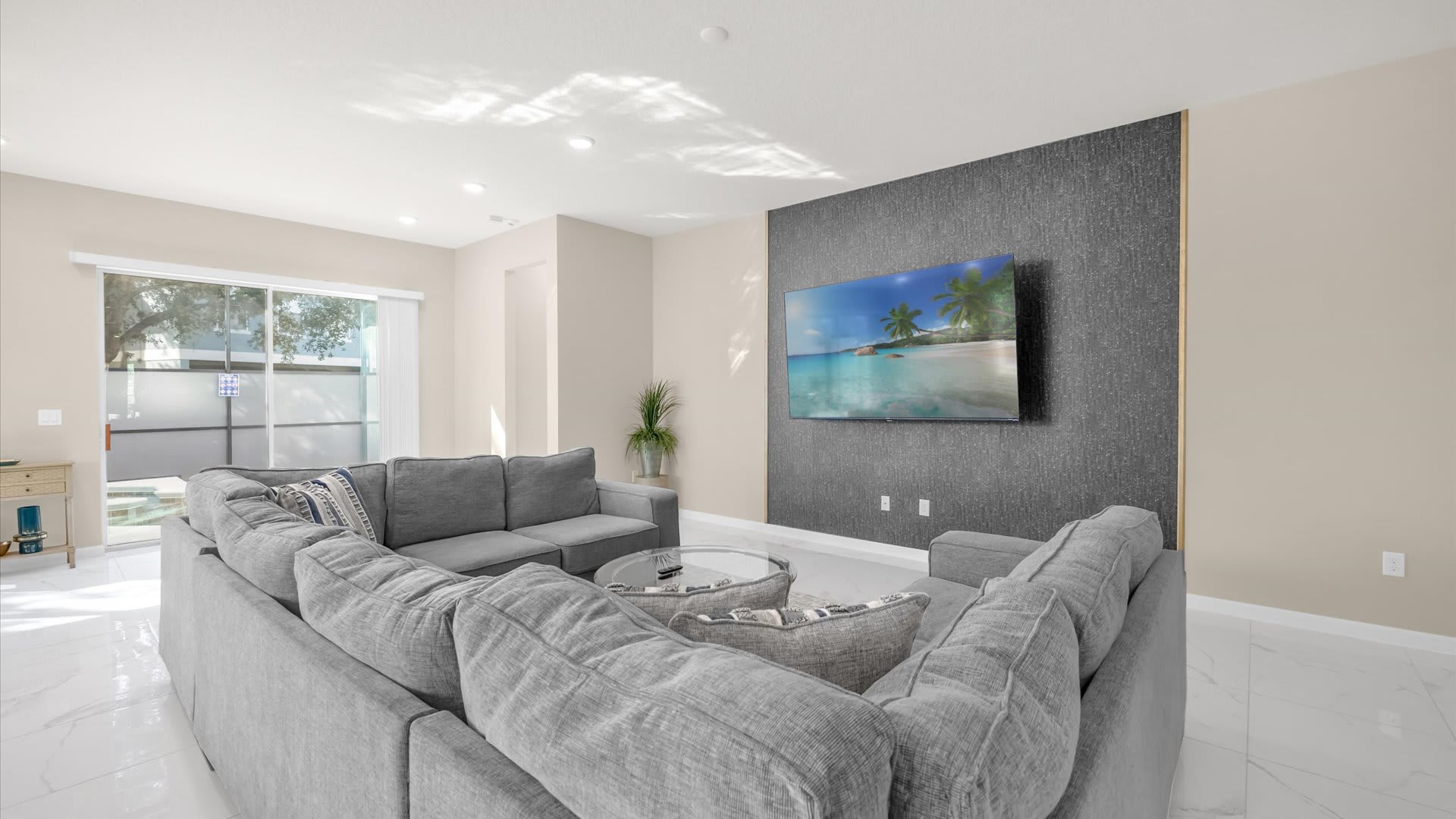 A bright and modern living room features a large gray sectional sofa on a patterned rug, facing a wall-mounted TV on a dark textured accent wall. Sliding glass doors lead to the outdoor pool area, and recessed lighting illuminates the space, which has light-colored walls and a glossy tile floor.