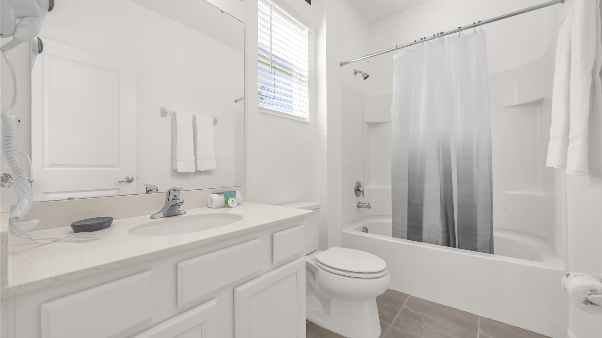 A clean, white bathroom featuring a single vanity with a light countertop, a toilet, and a shower/tub combo with a gray and white shower curtain. A window with blinds provides natural light.