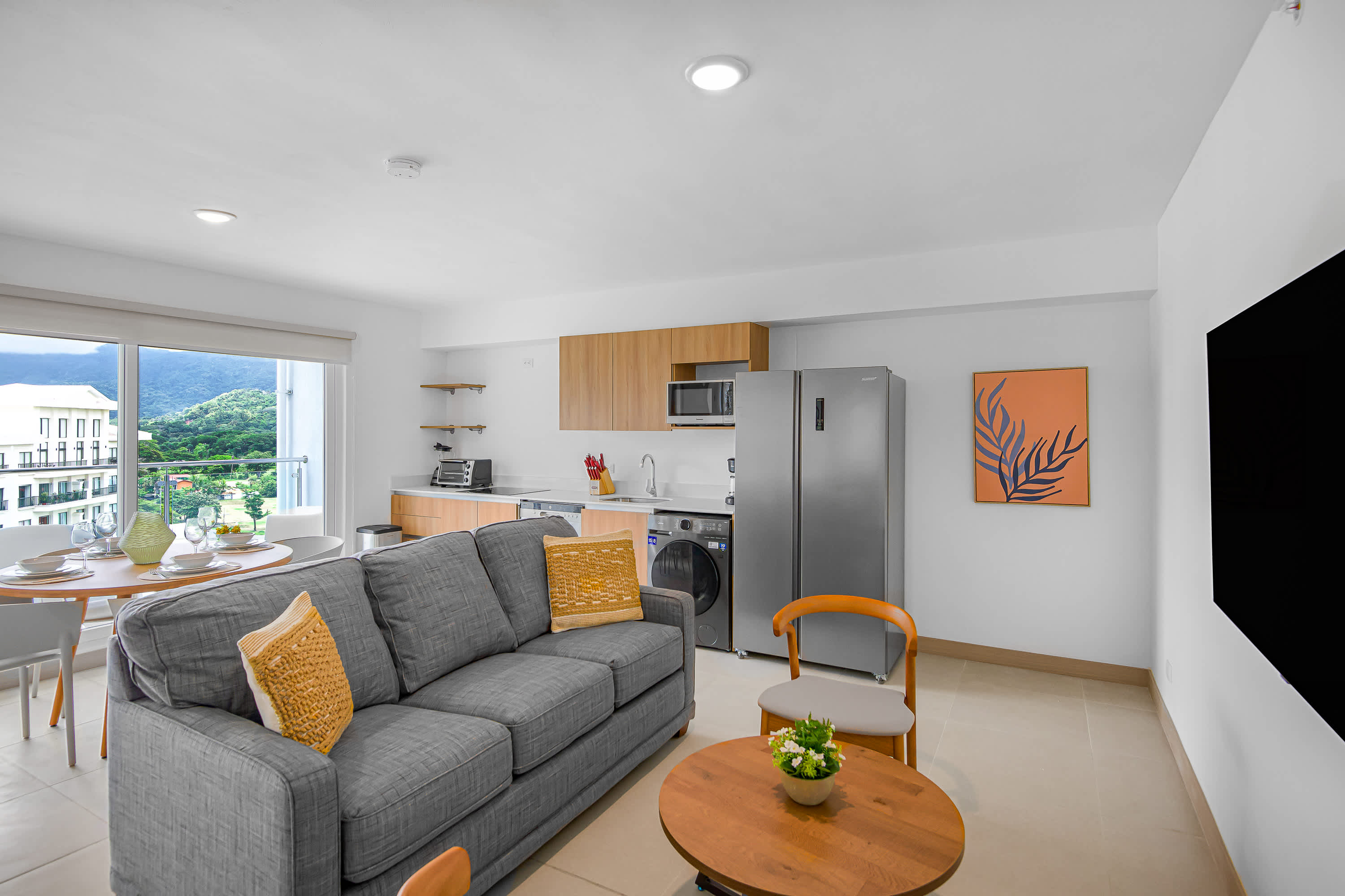 Modern open-concept living space with a gray sofa adorned with yellow pillows, a round wooden coffee table, and a dining area by a large window. The kitchen with light wood cabinetry and stainless steel appliances is visible on the right.